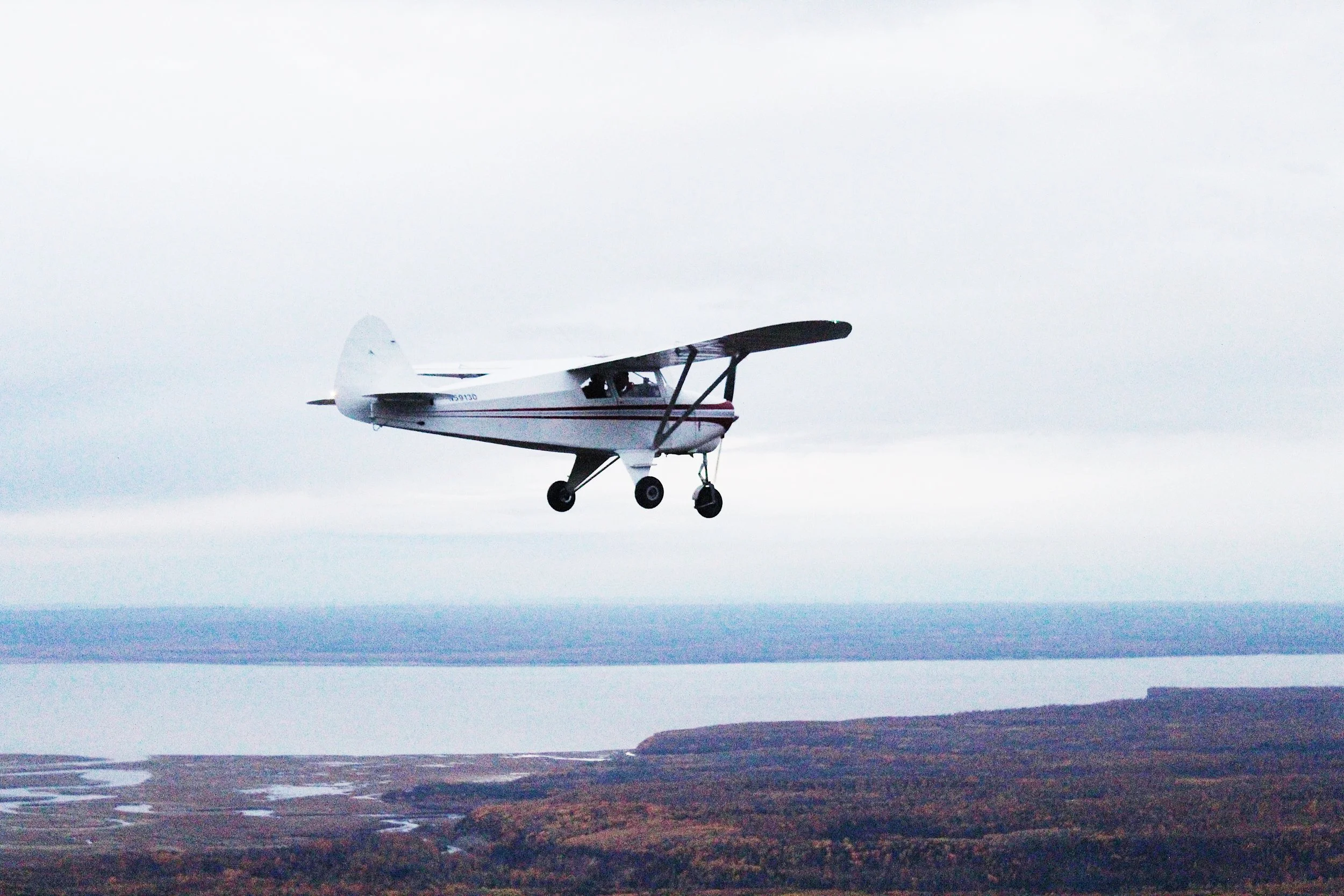 A small single-engine propeller airplane flying over a landscape with water and land, under an overcast sky.