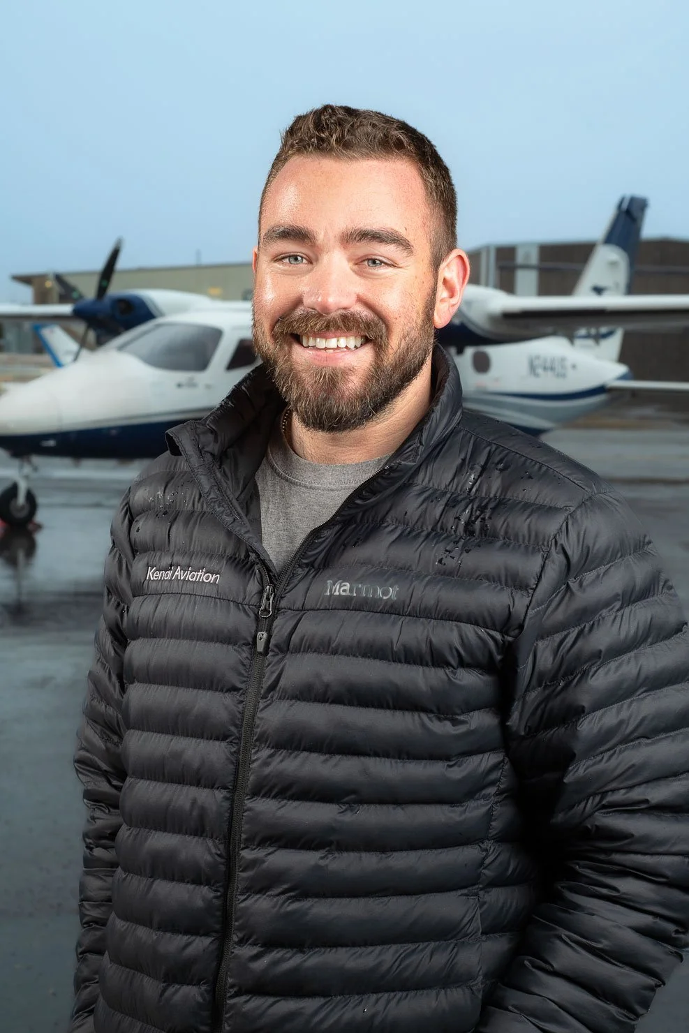A smiling man with a beard and short hair standing in front of small aircrafts at an airport, wearing a black puffy jacket.
