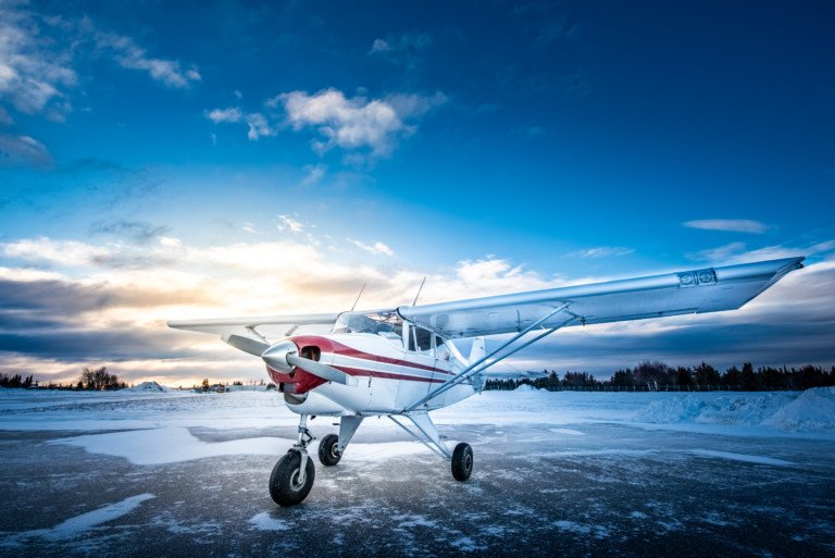 Small white airplane with red stripes parked on a snow-covered airstrip at sunset or sunrise under a partly cloudy sky.