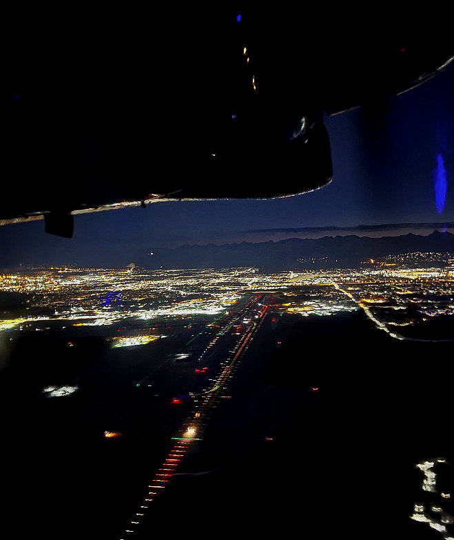 Nighttime view from an airplane window showing a brightly lit cityscape with an illuminated runway and mountains in the distance.