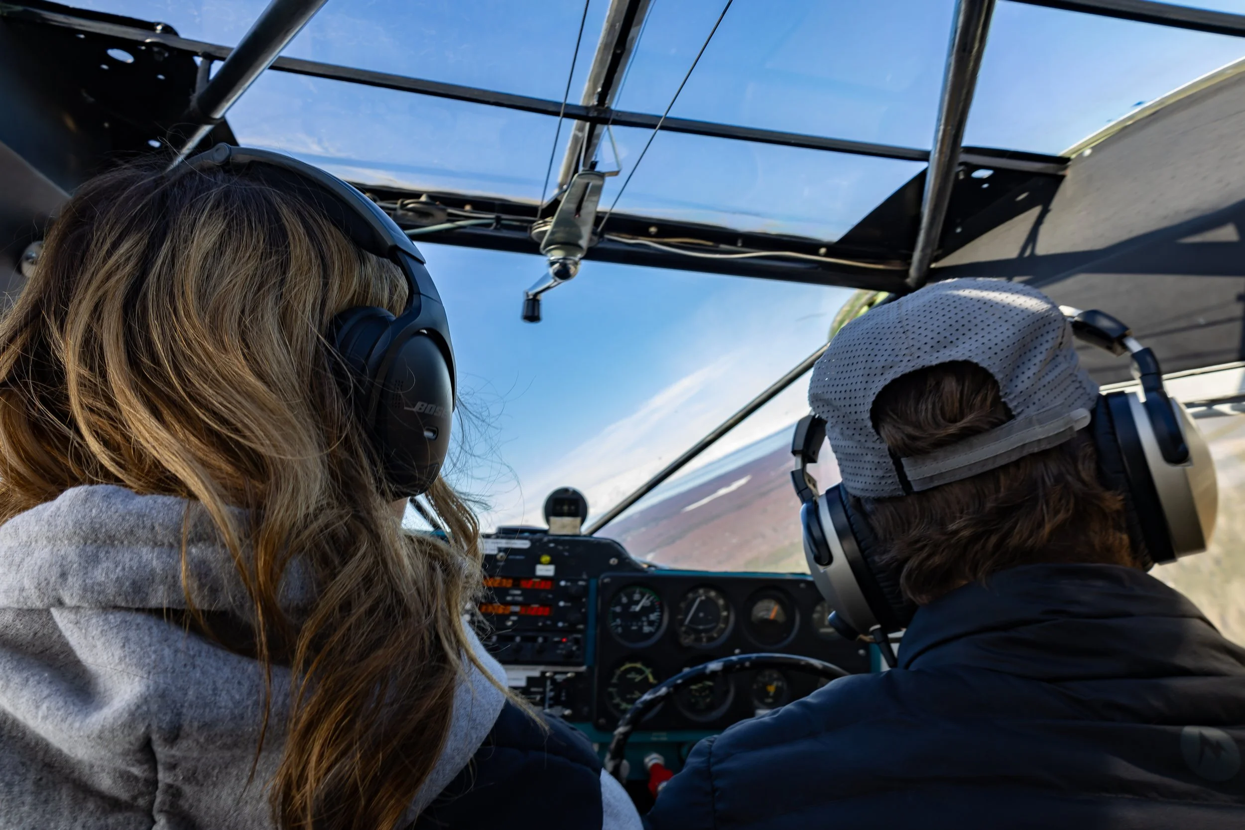 Two people flying in a small airplane, viewed from behind, with a view of the sky and landscape through the cockpit window.