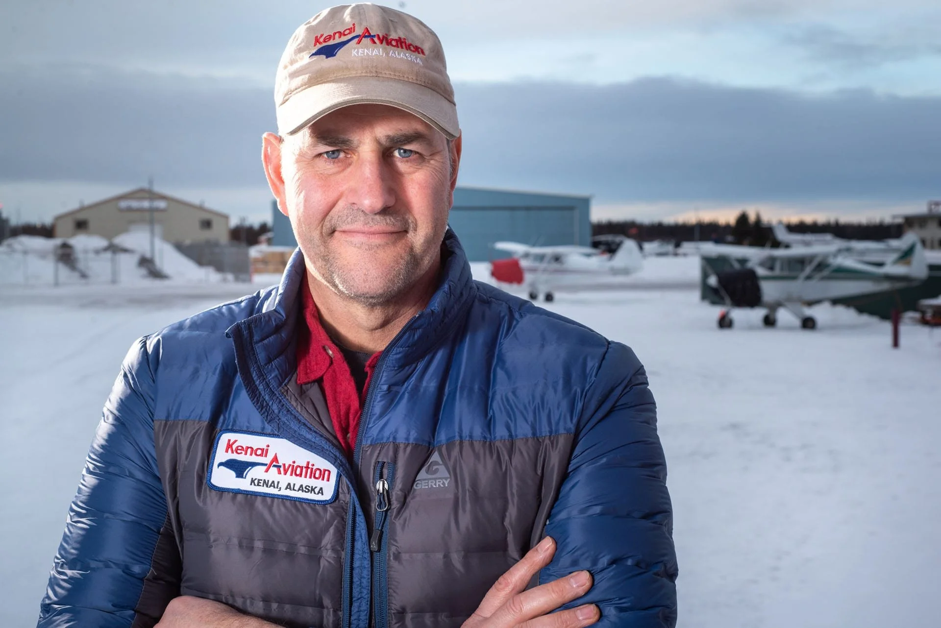 A man stands outdoors in a snowy landscape with small airplanes in the background. He wears a beige cap and a blue and black jacket with a patch that reads 'Kenai Aviation Kenai, Alaska.'