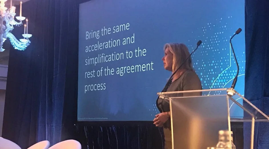 A woman stands at a clear lectern on a stage with a large screen behind her. The screen displays the text: 'Bring the same acceleration and simplification to the rest of the agreement process.' Blue curtains are in the background, and a chandelier hangs from the ceiling on the left side of the image.