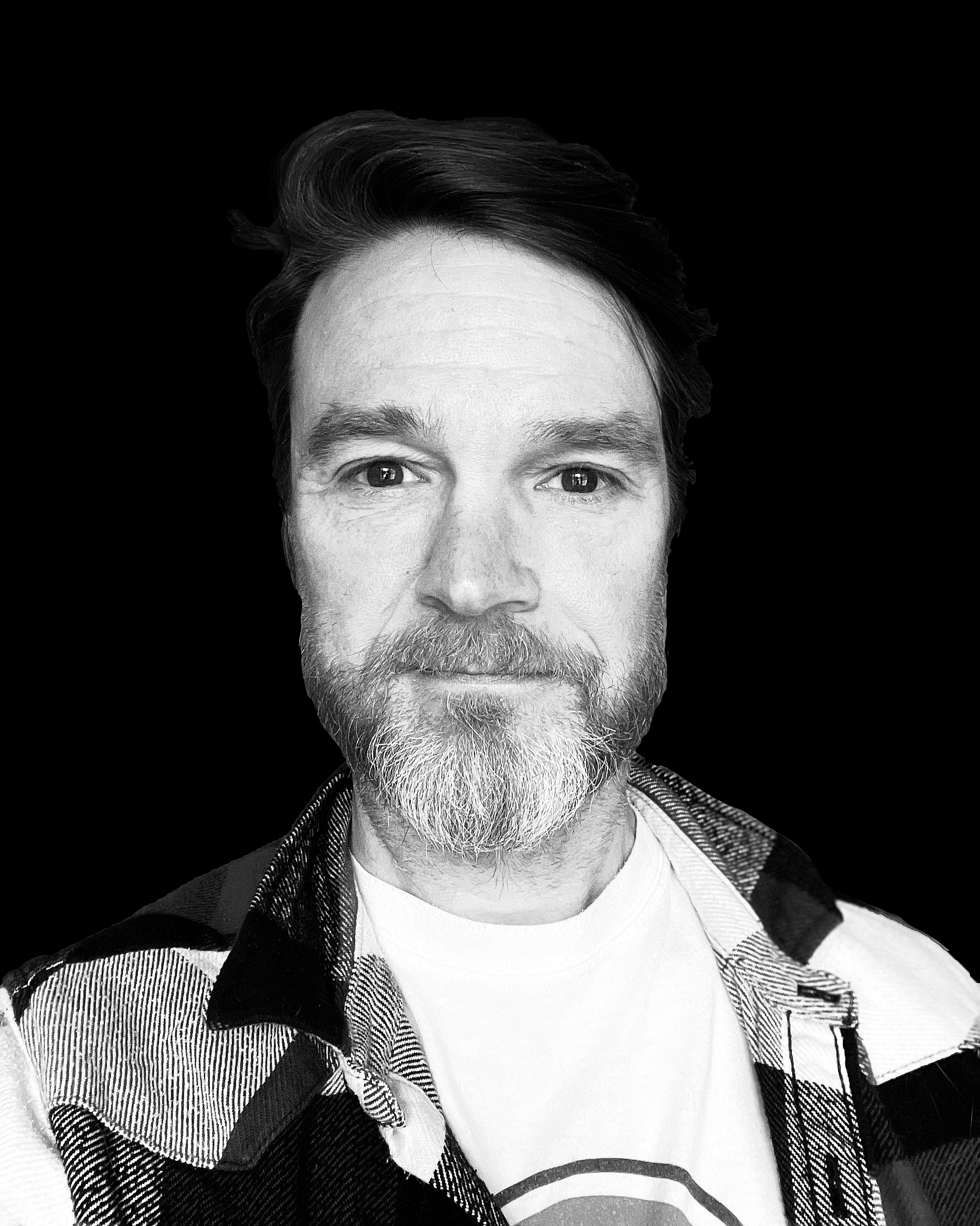 Black and white close-up portrait of a man with a beard and mustache, wearing a plaid shirt and a t-shirt with a circular logo, against a solid black background.