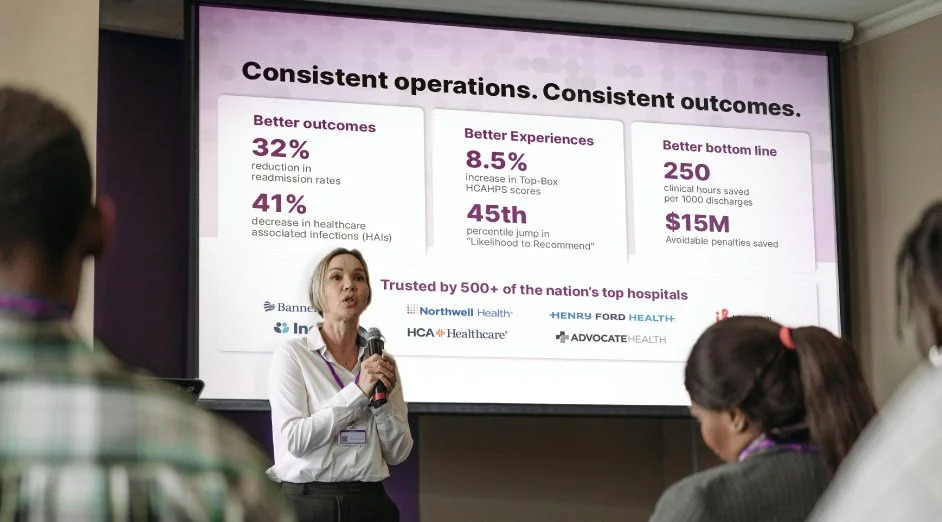 A woman giving a presentation on healthcare metrics to an audience, with a slide displaying statistics such as improved outcomes, better experiences, and healthcare savings.