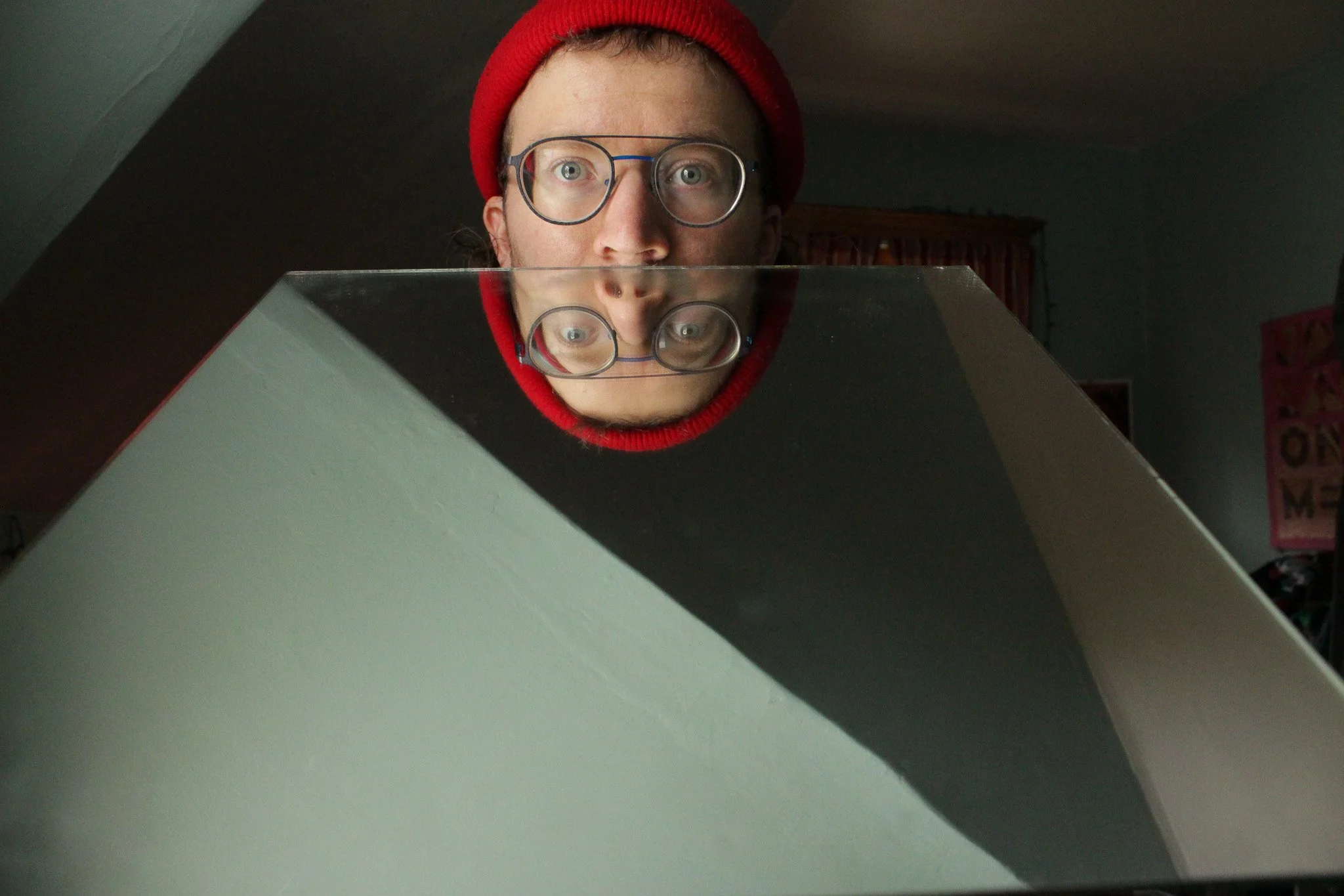A person with glasses and a red beanie looking into a reflective glass surface, creating a mirror image of their face.