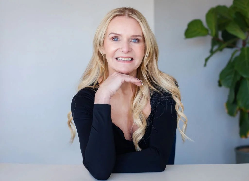 A blonde woman with blue eyes sitting at a table, smiling and resting her chin on her hand, in front of a light-colored background with a large green plant on the right.