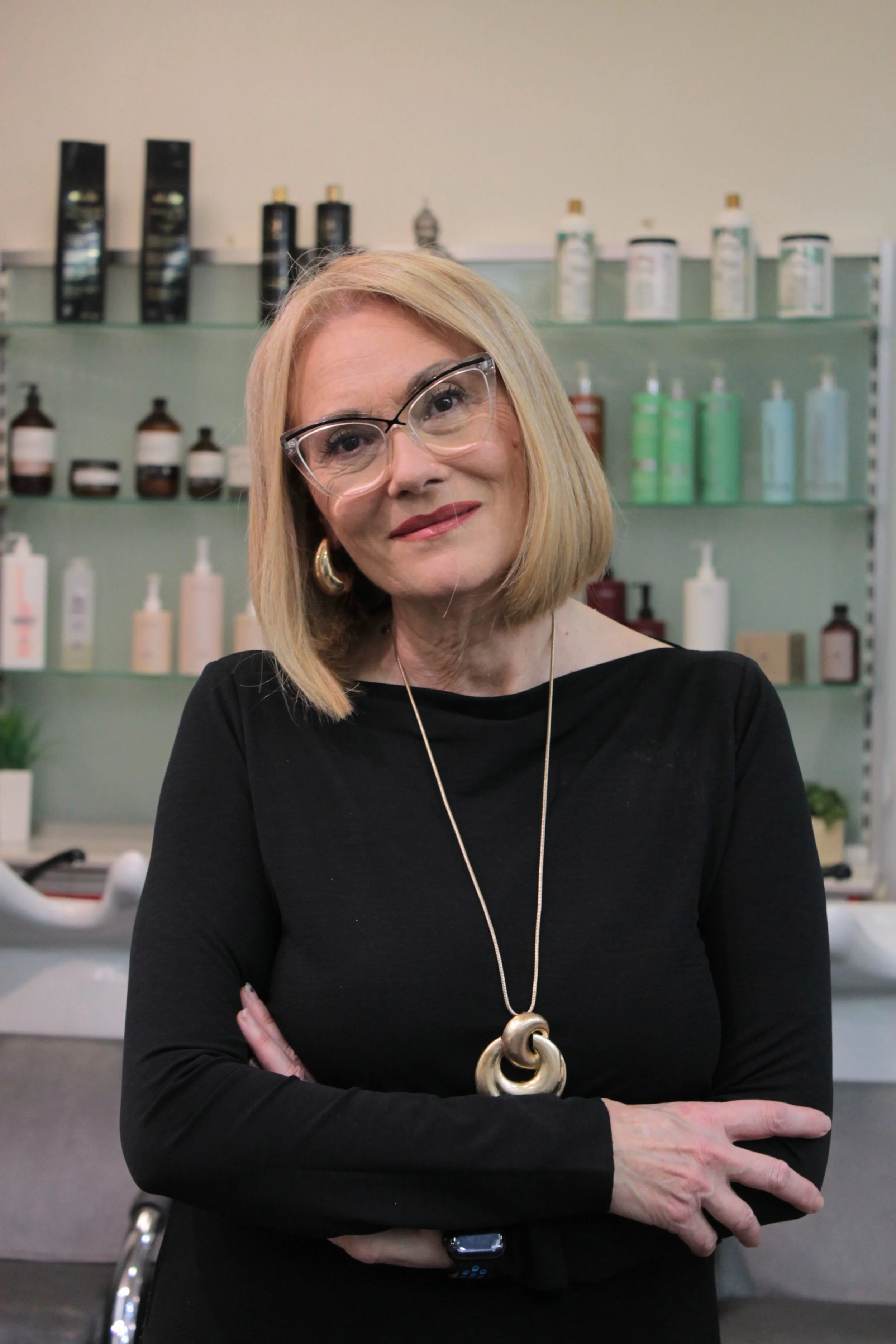 A woman with blonde hair, glasses, and gold jewelry standing in front of shelves with hair and skincare products.