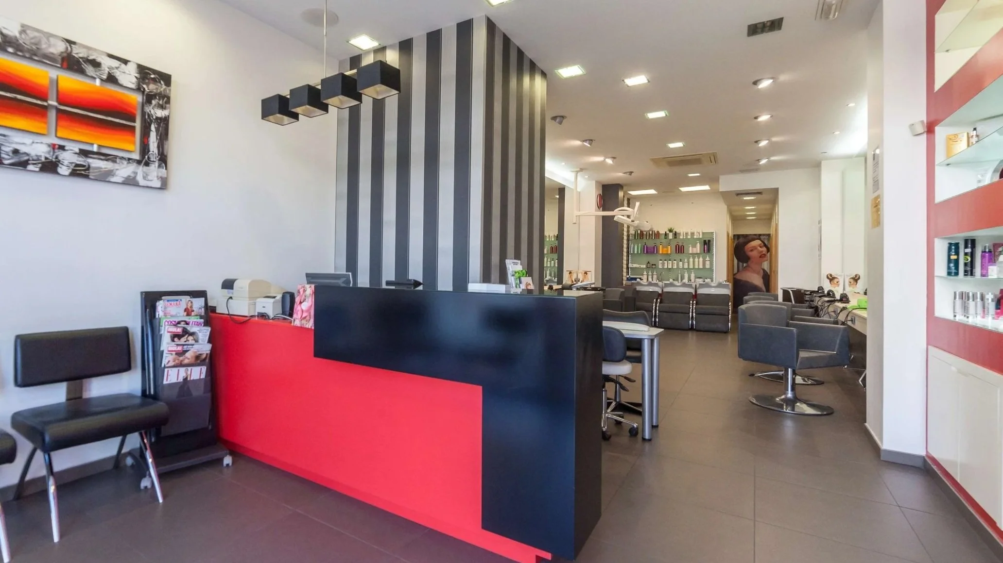 Interior of a modern hair salon with black and gray chairs, a black and red reception desk, shelves with hair products, and a wall with salon services advertisement.
