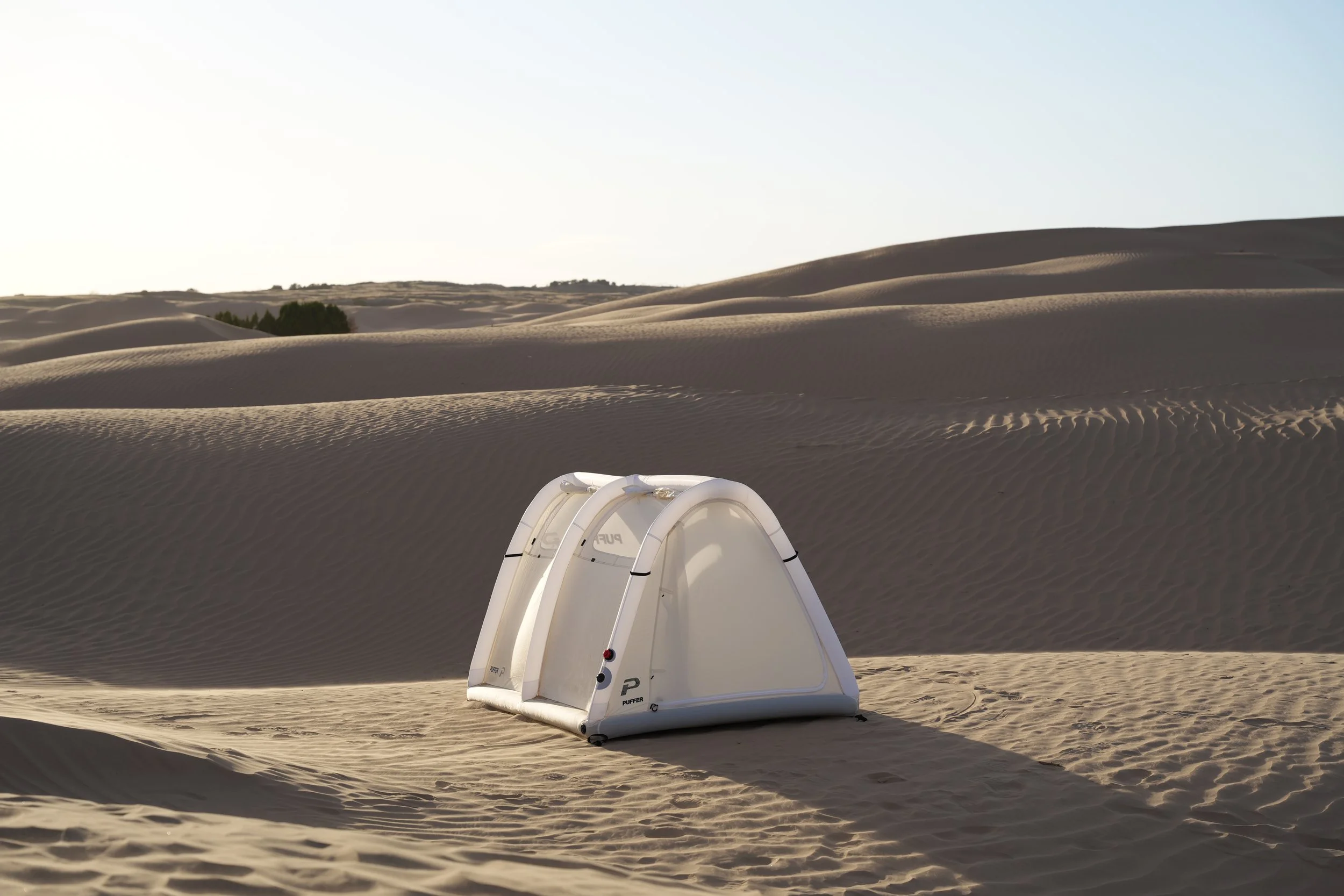 A desert landscape with sand dunes and a white portable inflatable tent  in the foreground.
