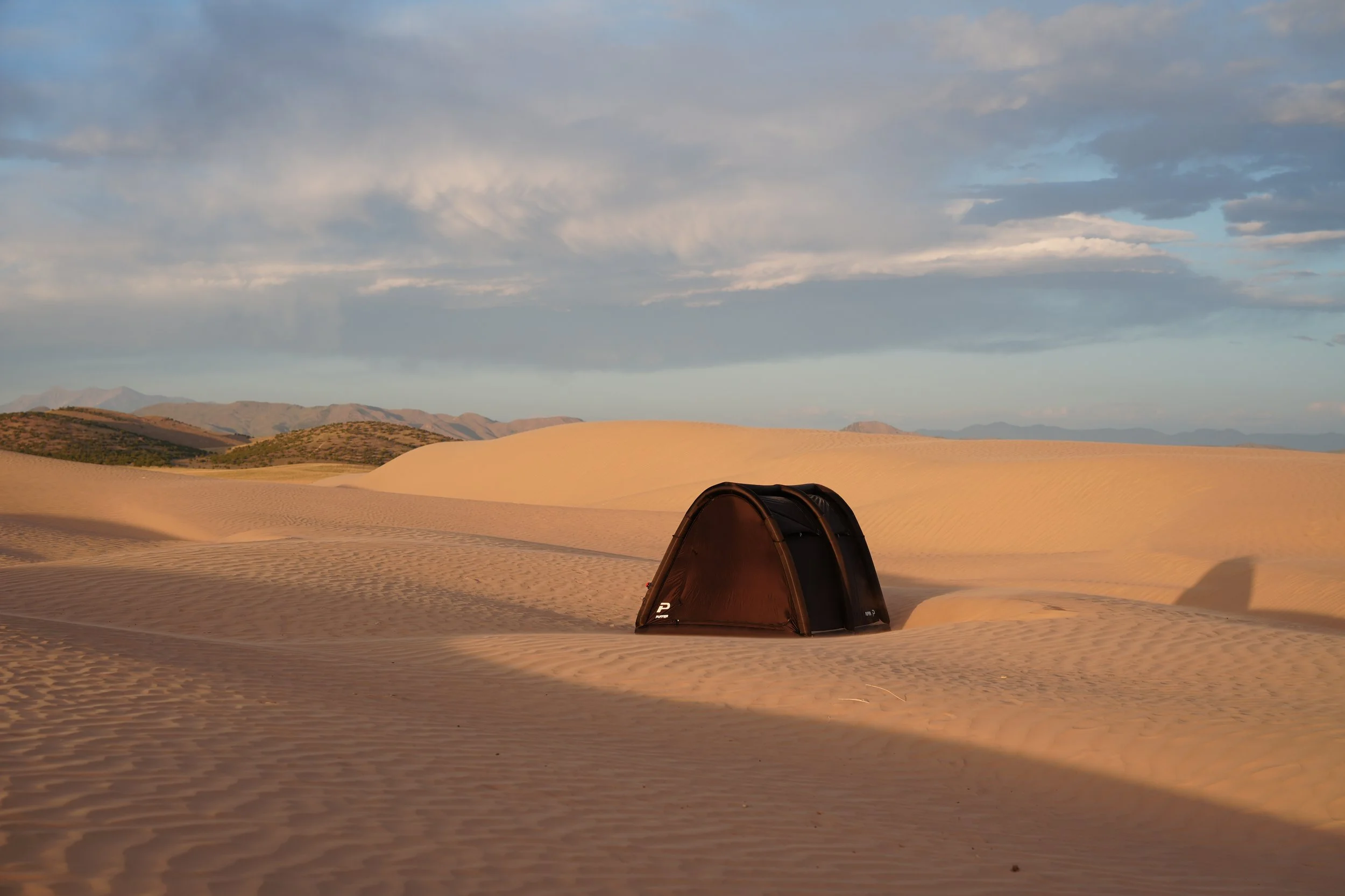 A desert landscape with sand dunes, some distant mountains, and a cloudy sky. There is a small, dark tent partially in shadow in the foreground.