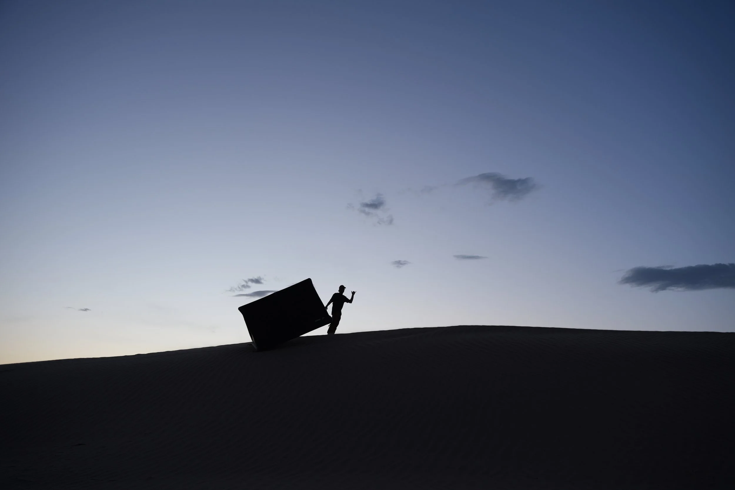 Silhouette of a person pulling a puffer tent on a sand dune during twilight with a darkening sky and scattered clouds.