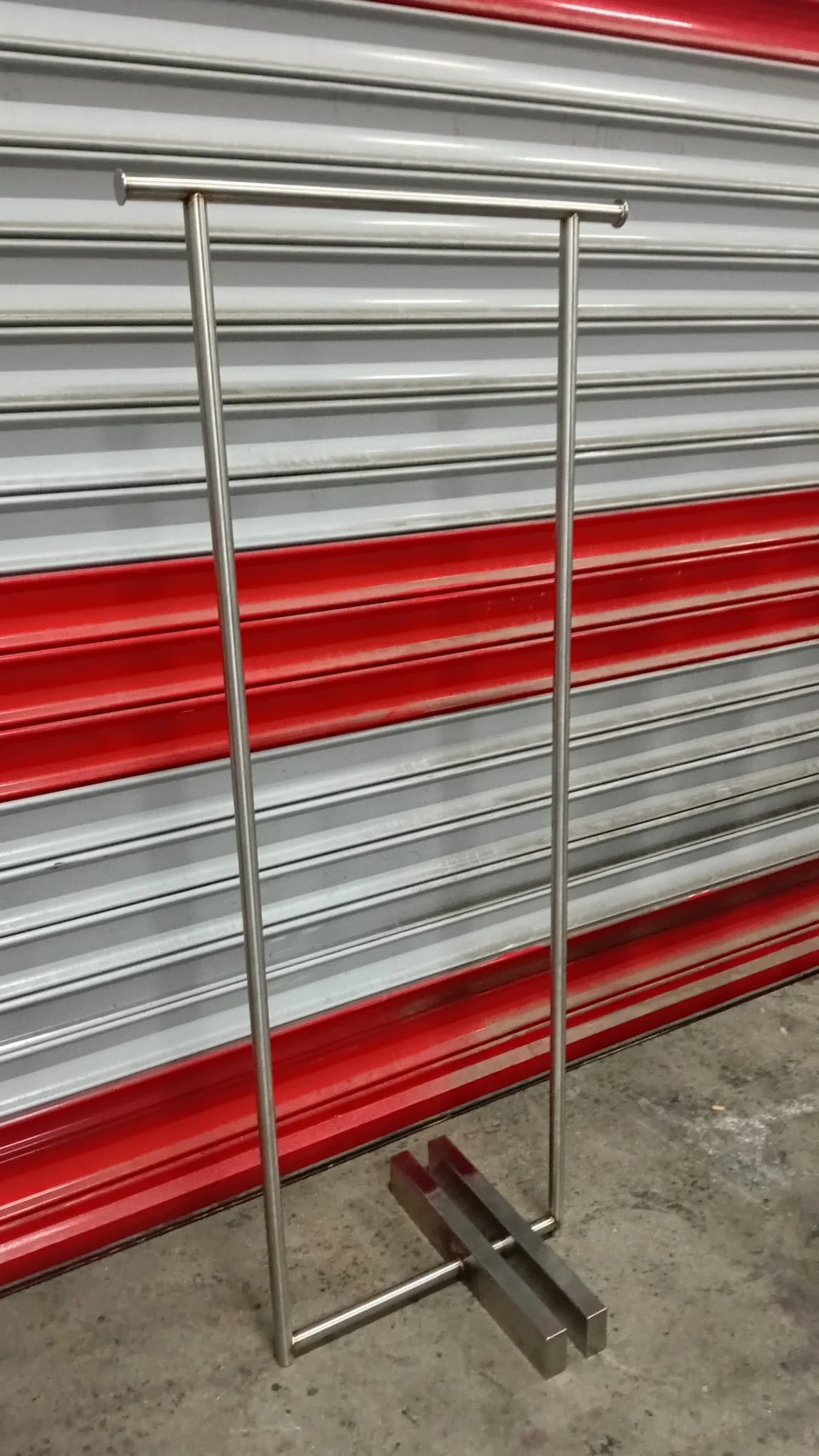 Pilates veya yoga aleti olan bir metal dik duruş çubuğu, arka planda kapalı olan bir mağaza veya depo kapısının kırmızı ve gri renkli metal panelleri.