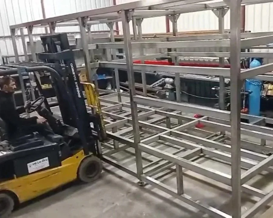 A person operating a yellow and black forklift machine in a large industrial warehouse with metal shelving and equipment.