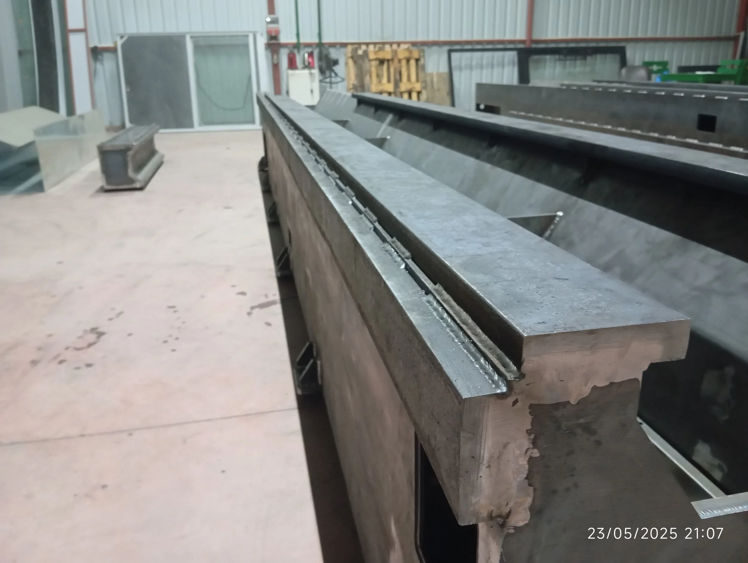 Endüstriyel bir ortamda büyük bir metal parça yer almakta, arka planda ise çeşitli aletler ve malzemeler var. Çalışma alanı beton zemin üzerinde ve iç mekân görünümünde.