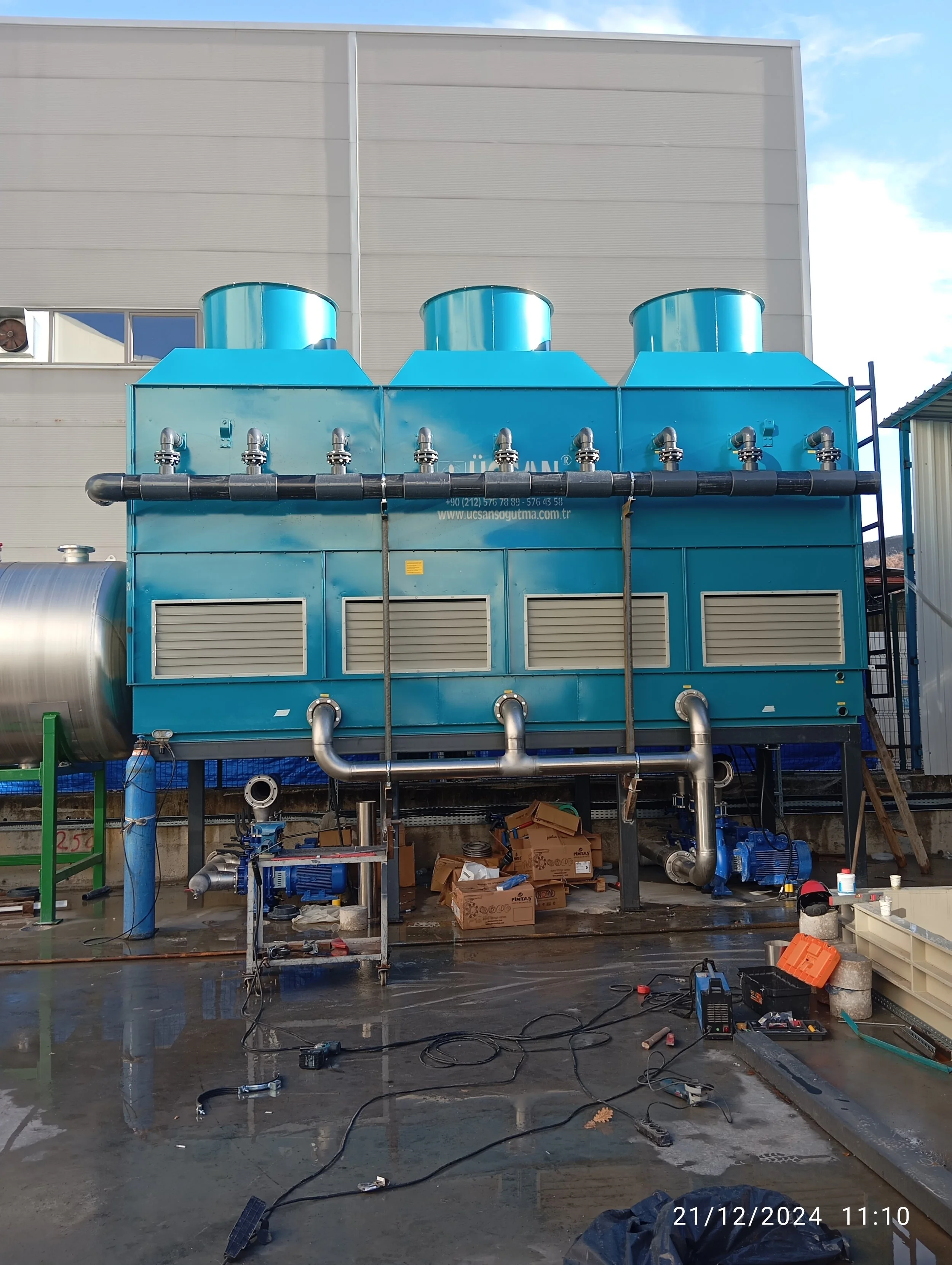 Endüstriyel soğutma veya klima sistemi, büyük mavi yapılar ve borularla çevrelenmiş, çevrede araç ve ekipmanlar bulunuyor.