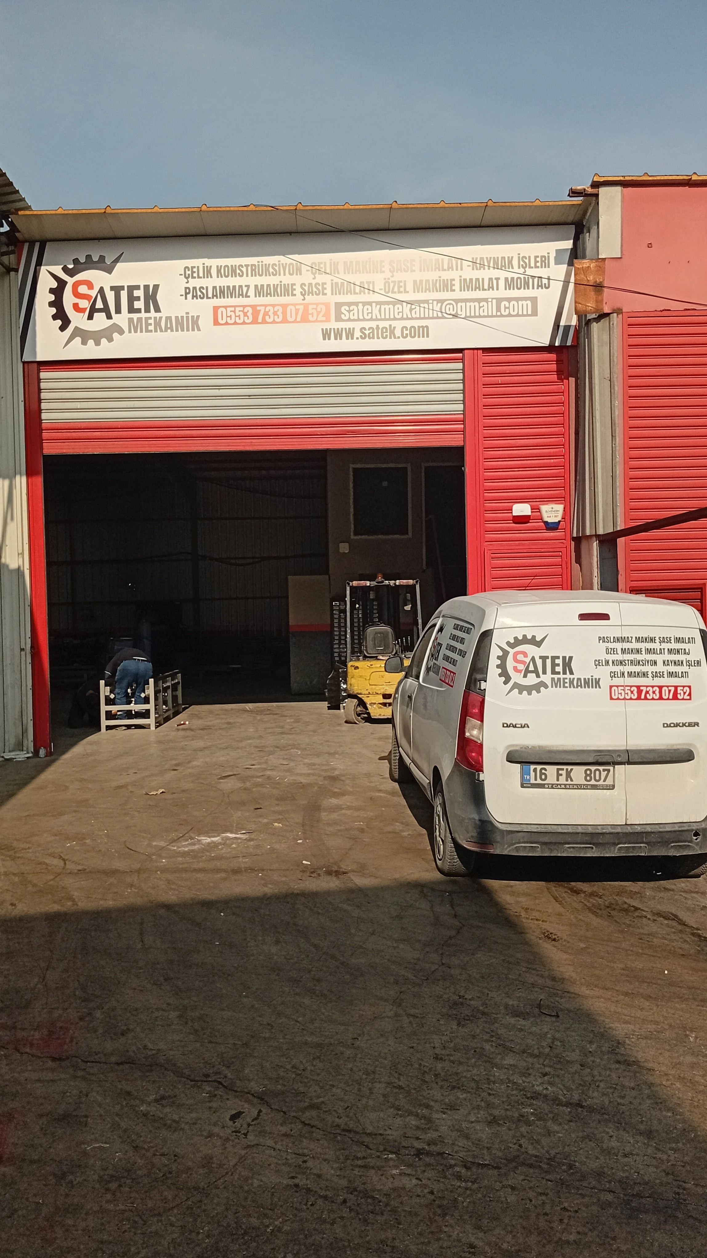Photo of a workshop with a large red rolling shutter door partially open. A white van with the company's logo and contact information is parked in front. Inside, there is a person working near some equipment. A small forklift is present inside the workshop.