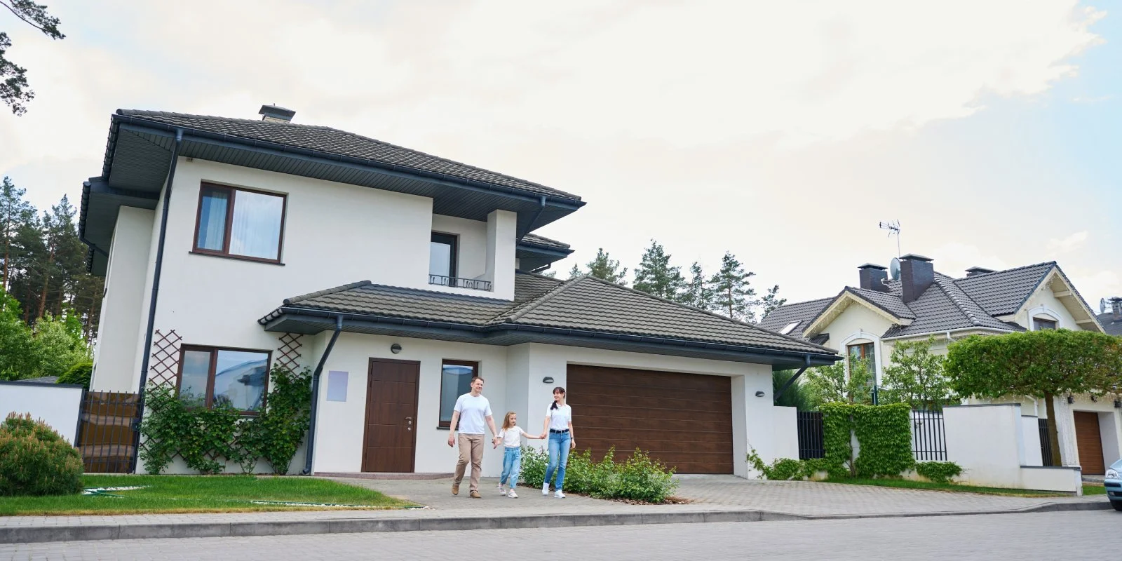 A family of three walking on the sidewalk in front of their modern house with a garage, white walls, brown doors and windows, and surrounded by lush greenery.