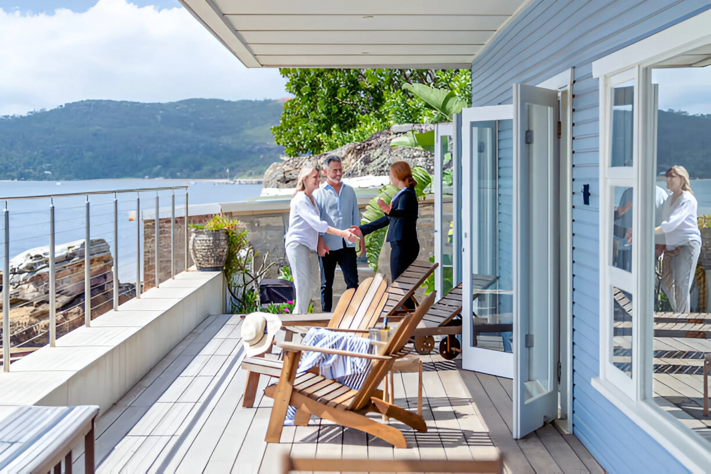 People shaking hands on a seaside deck with lounge chairs, a hat, and a striped towel, near a blue house with open windows, with water and mountains in the background.