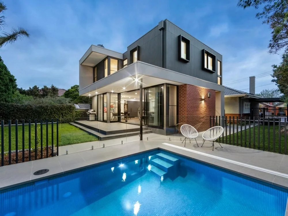 Modern two-story house with large windows, balcony, and an outdoor swimming pool with two white chairs nearby, surrounded by a black fence and green lawn, under a partly cloudy sky.