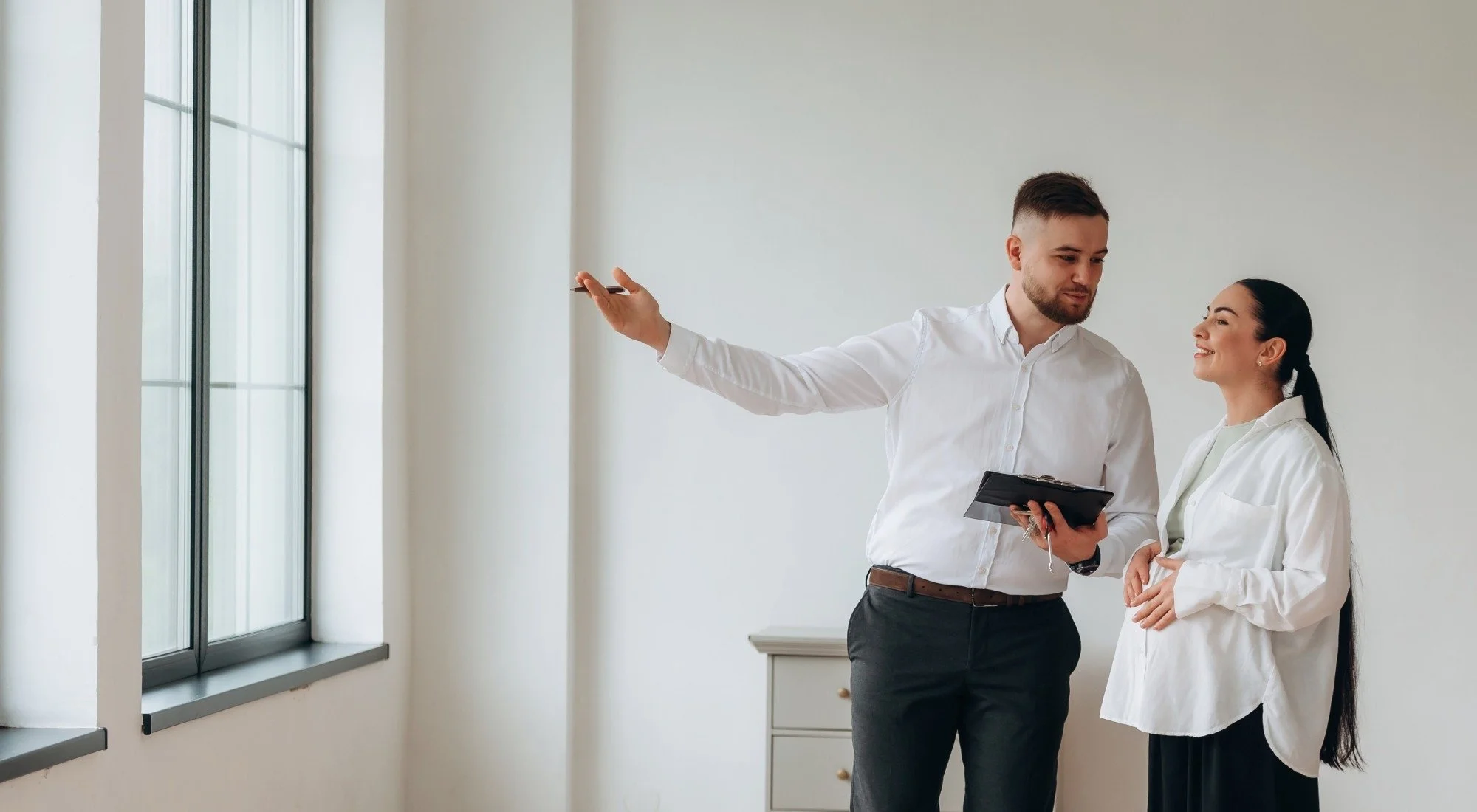 A man and woman, both dressed in white shirts, are having a conversation indoors near large windows. The man is gesturing with his left hand and holding a clipboard in his right hand. The woman is smiling and listening.