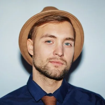 A young man with light brown hair, a beard, wearing a tan fedora, navy dress shirt, and a brown tie, posing against a plain blue background.