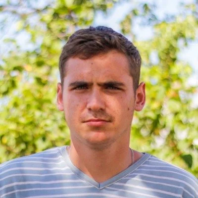 Young man with short brown hair and a serious expression, wearing a gray and white striped shirt, standing outdoors with green foliage in the background.
