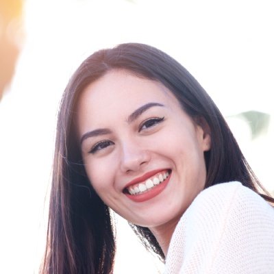 A young woman smiling, with long dark hair, wearing a white top, in front of a bright background.