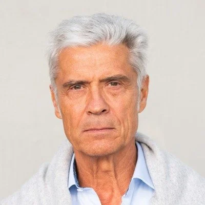 Close-up of an older man with white hair and serious expression, wearing a light blue shirt and a gray sweater, against a plain neutral background.