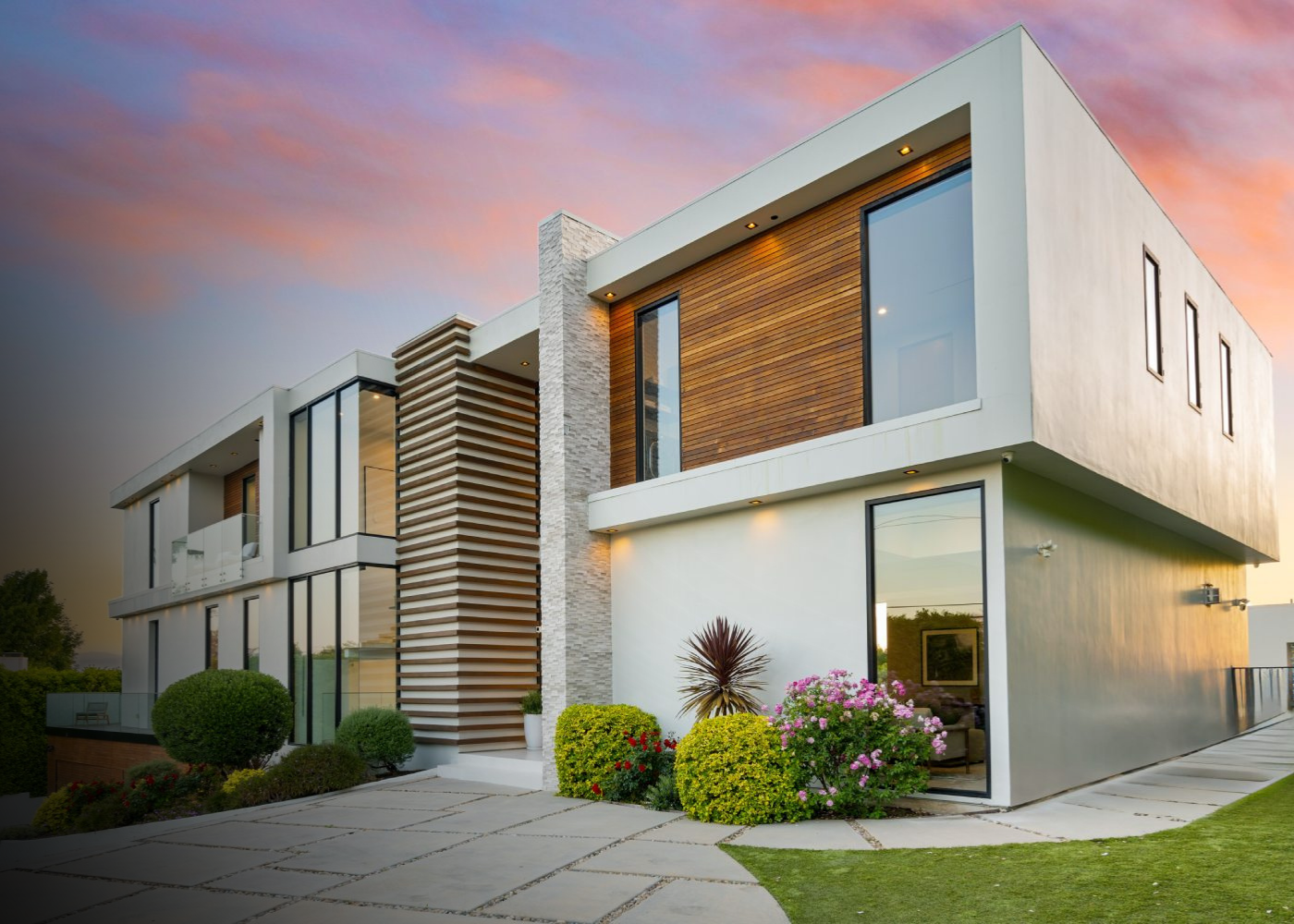 Modern multi-story house with large windows, white exterior walls, wood panel accents, and landscaped front yard with bushes and flowers during sunset.