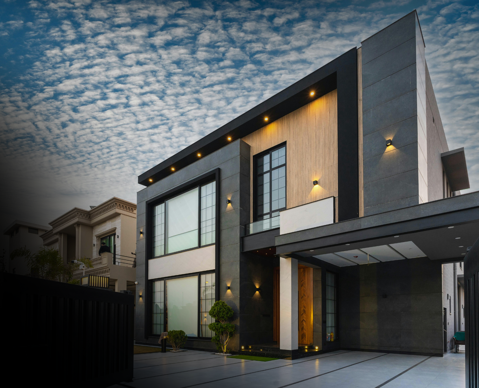 Modern two-story house with black and wooden exterior accents, large glass windows, and well-lit outdoor area, under a partly cloudy sky at dusk.