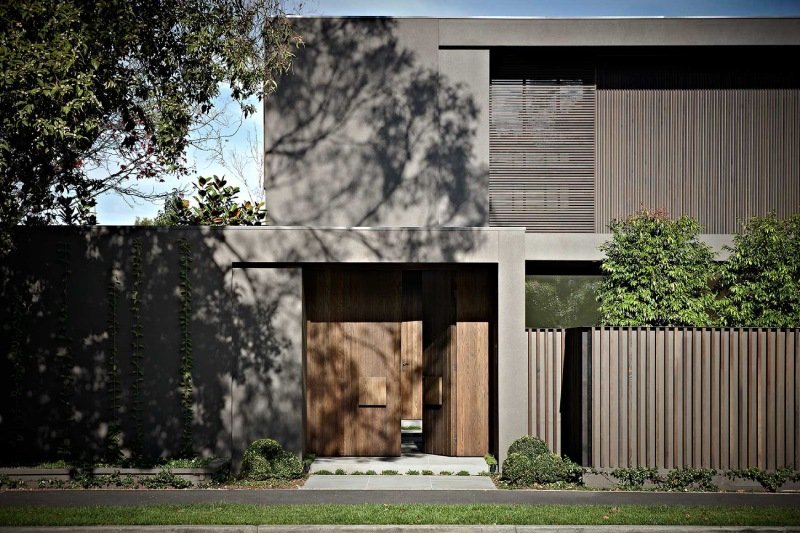 Modern house with wooden doors, concrete facade, and wooden fencing, surrounded by greenery and trees casting shadows.