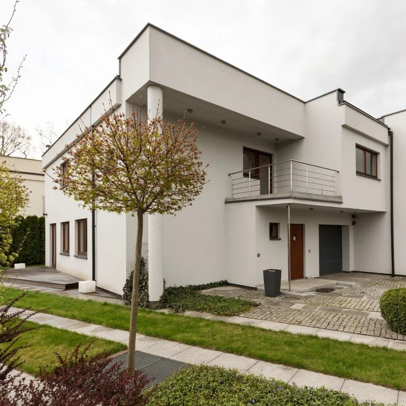 Modern white two-story house with a balcony, front door, driveway, and manicured lawn with trees and bushes.