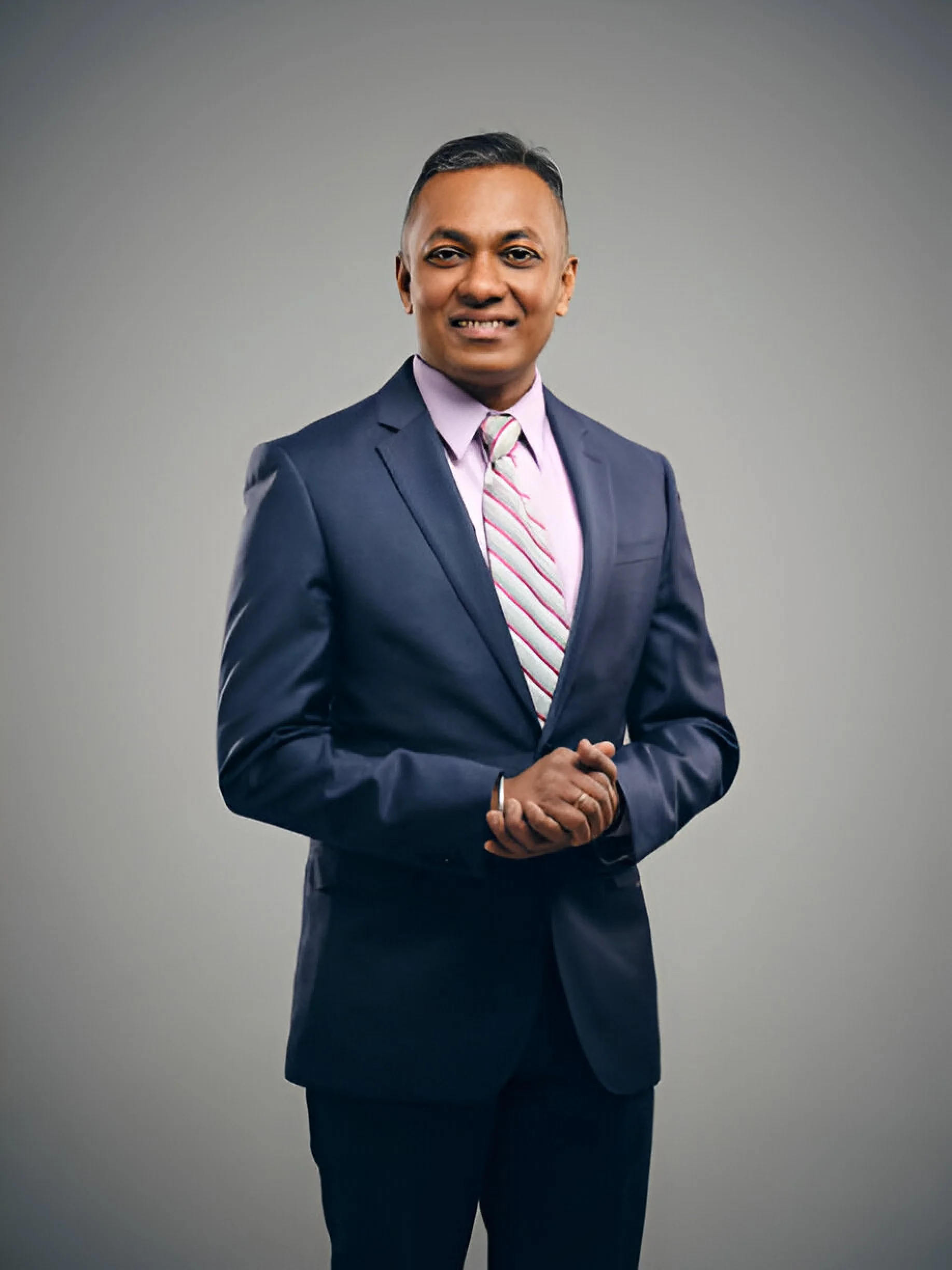 A professional man in a navy suit, pink shirt, and striped tie standing against a gray background.
