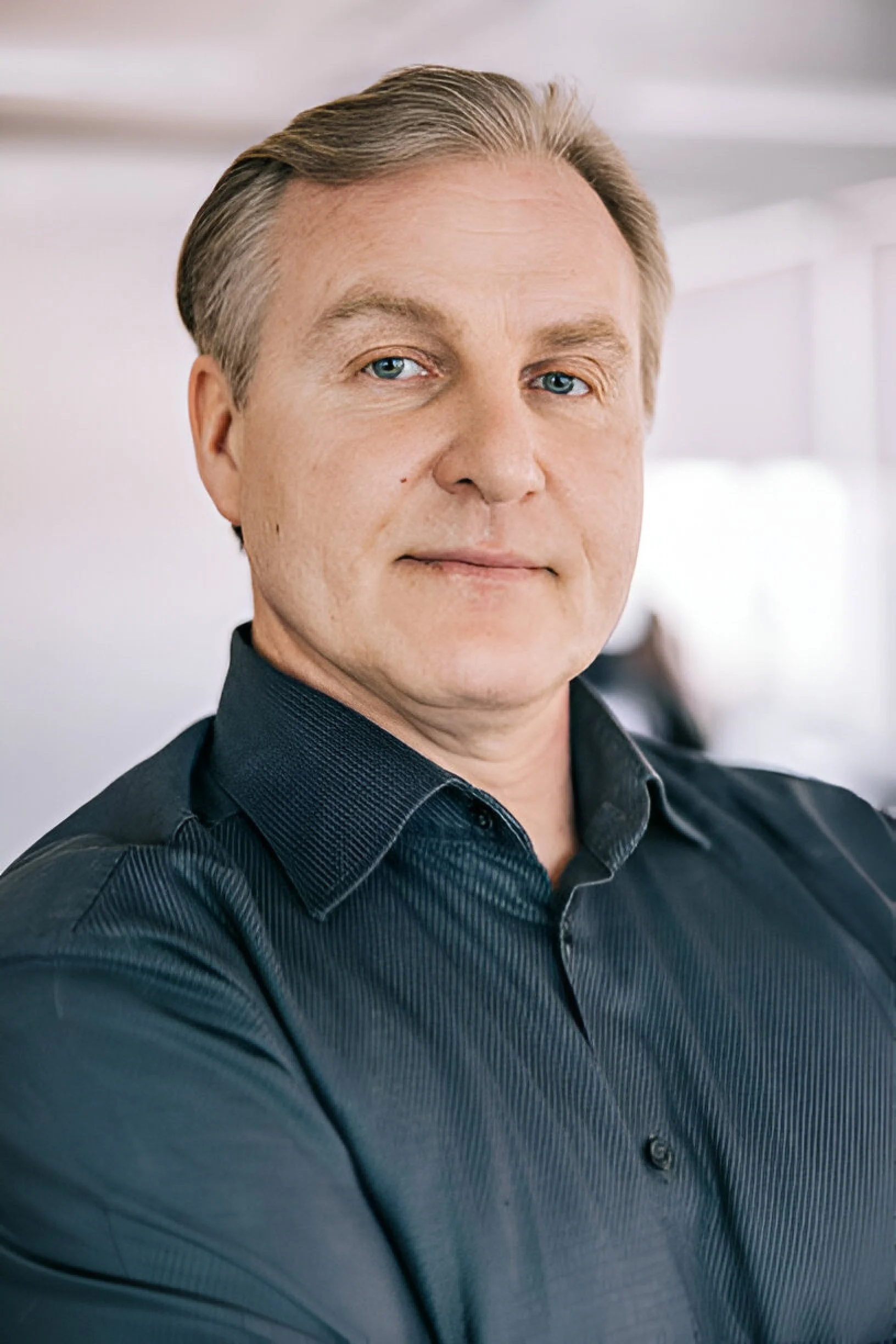 A man with blonde hair and blue eyes wearing a dark blue dress shirt, posing indoors with a blurred background.