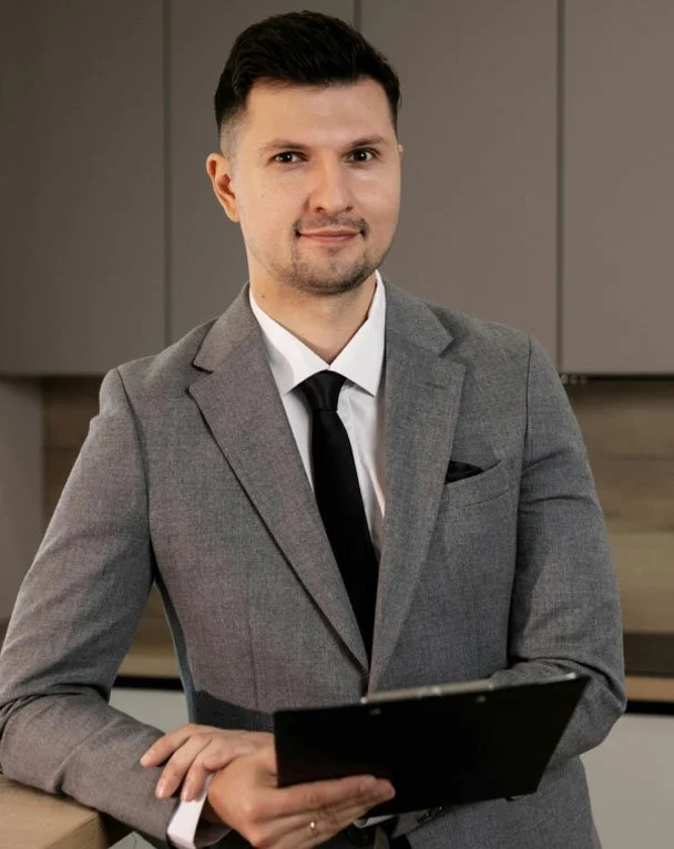 A man dressed in a gray suit with a black tie and white shirt, holding a black clipboard, standing in a modern office setting.
