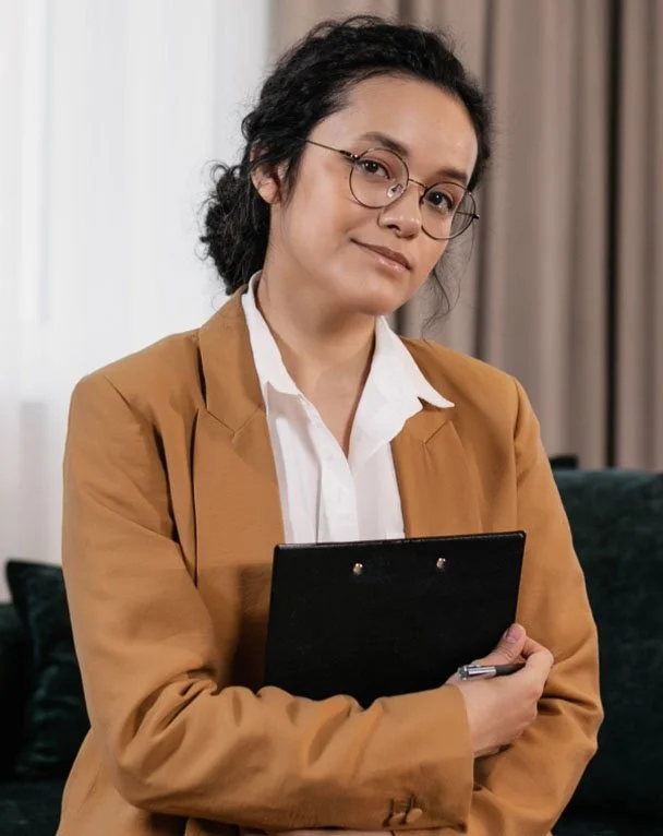 A woman with glasses, dark curly hair, wearing a white blouse and a tan blazer, holding a black clipboard, standing indoors with a neutral expression.