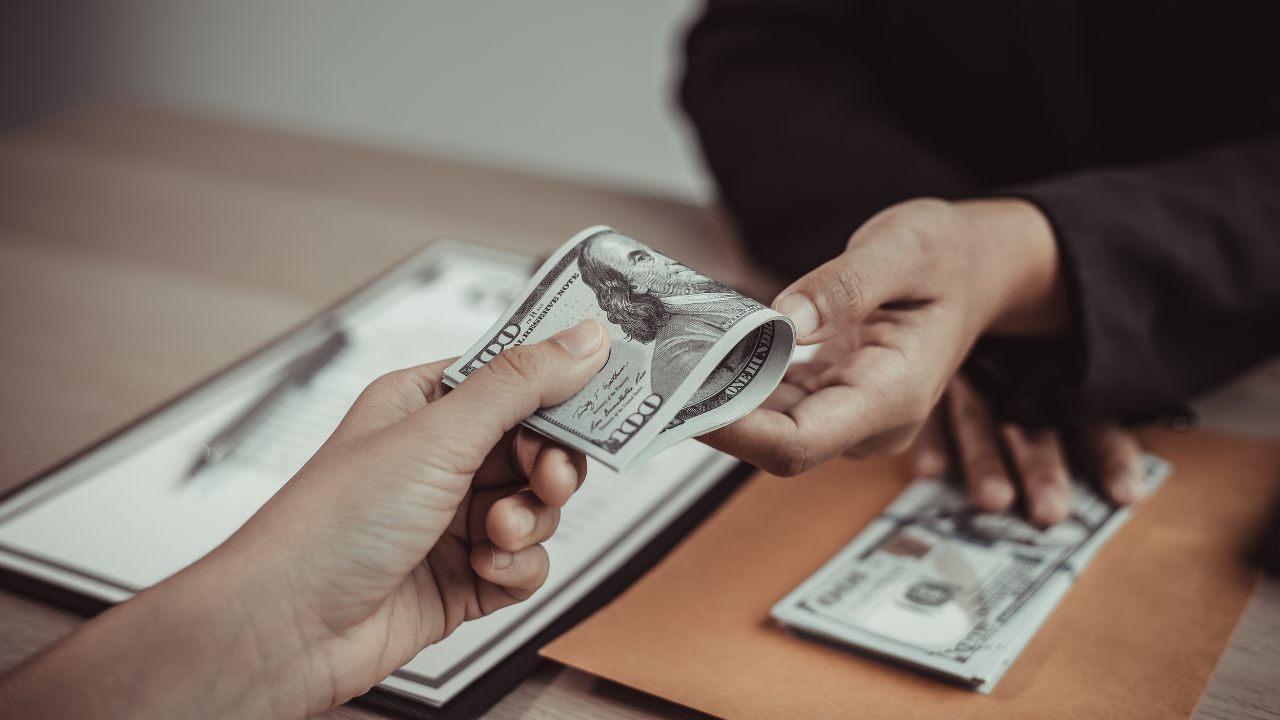 Two people exchanging hundred-dollar bills across a desk in an indoor setting.