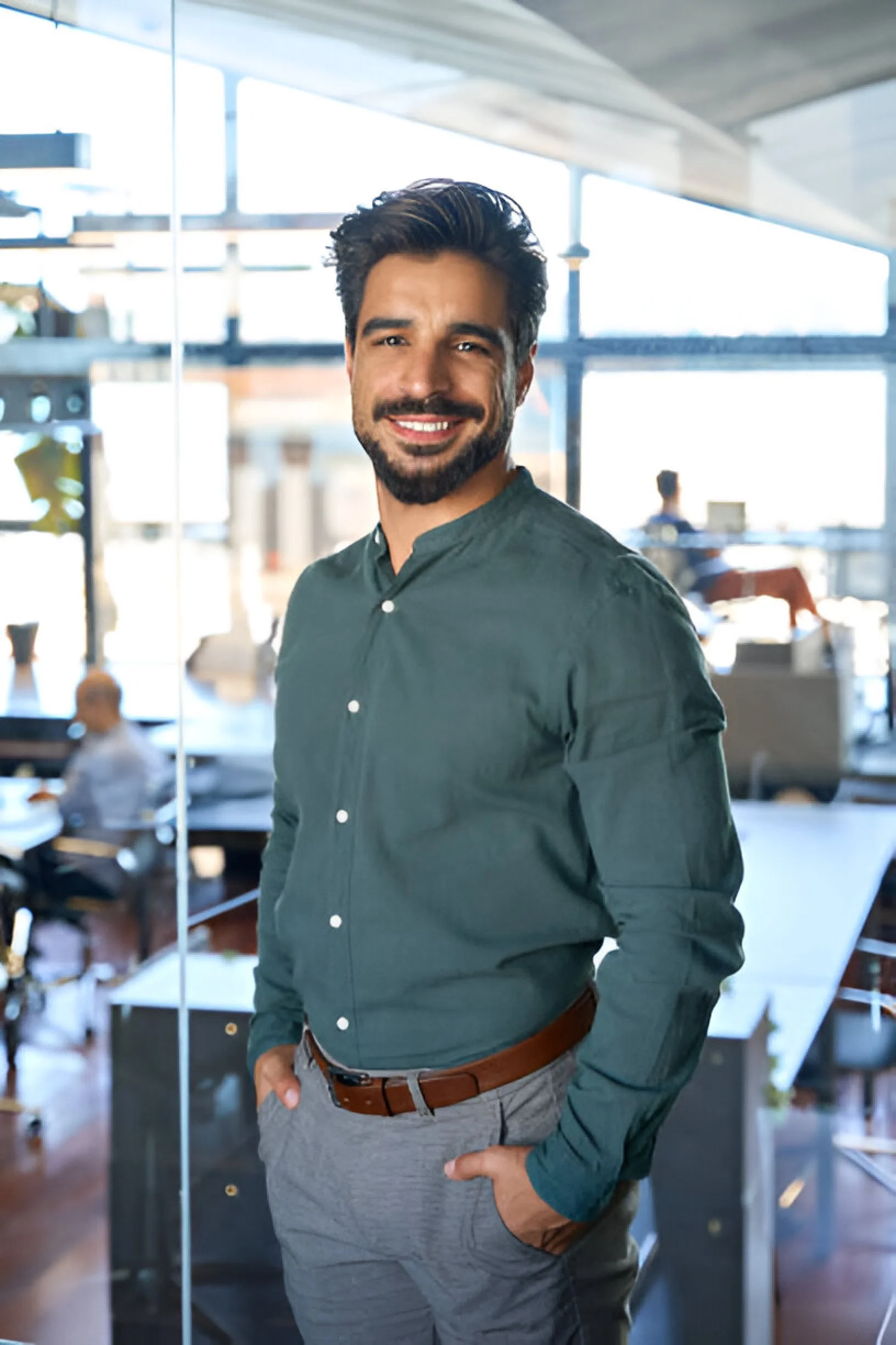 A man with dark hair and a beard, smiling and posing with his hands in his pockets in a modern office space with large windows, a glass wall, and people working in the background.