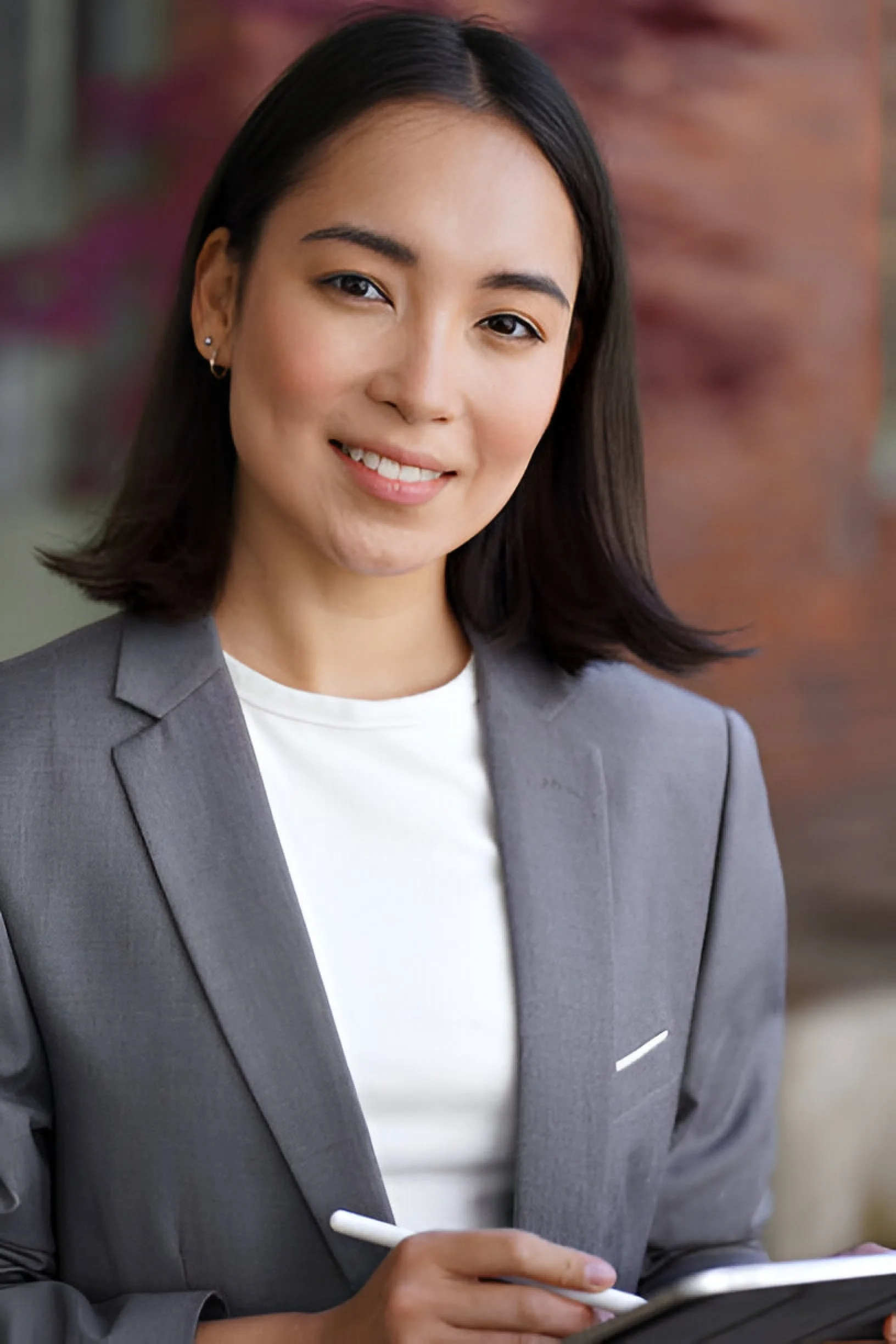 A woman with shoulder-length dark hair wearing a gray blazer and white shirt, smiling, holding a stylus and a tablet, in an urban background.