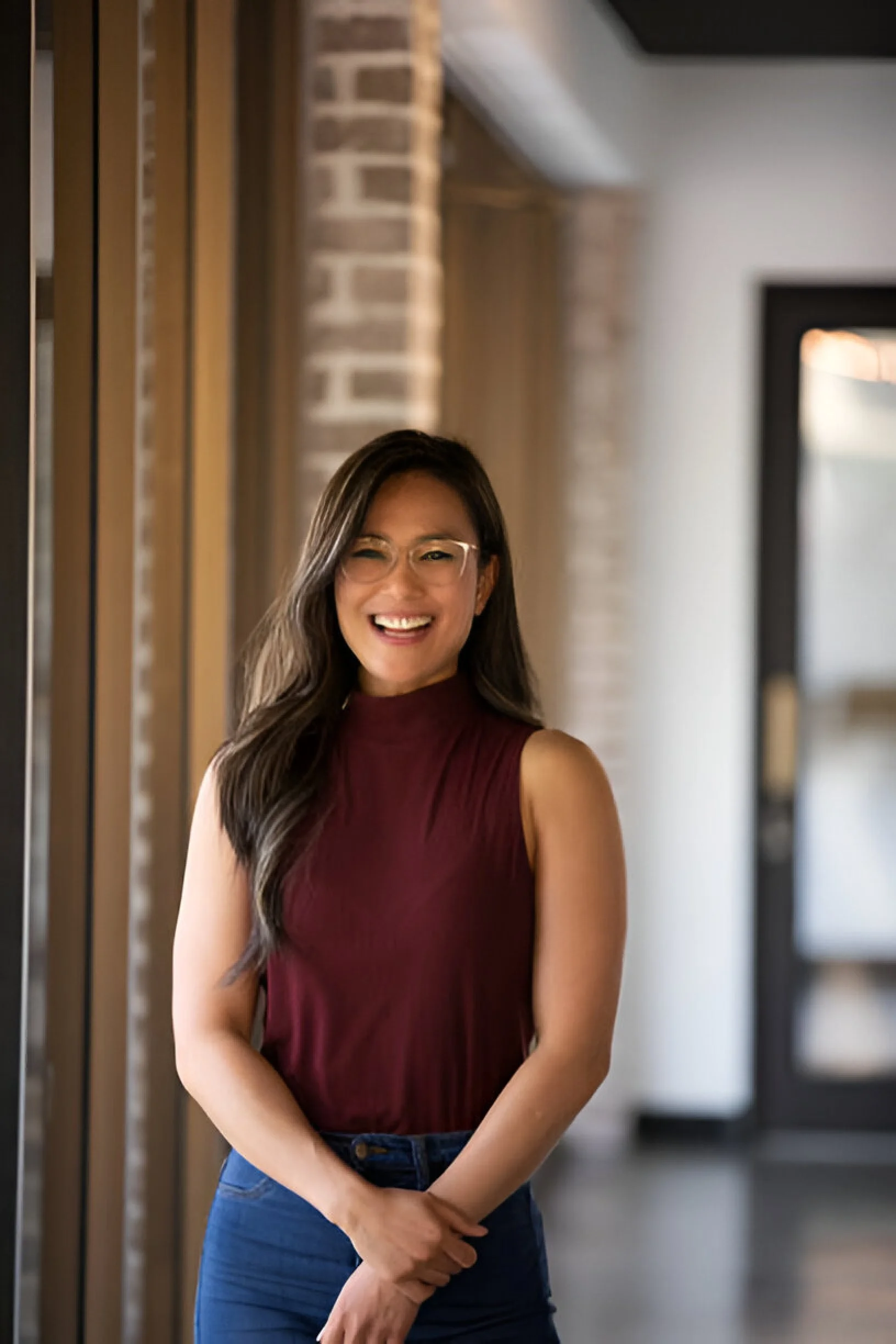 A woman with long dark hair and glasses smiling and standing in a hallway with wooden walls and brick accents.