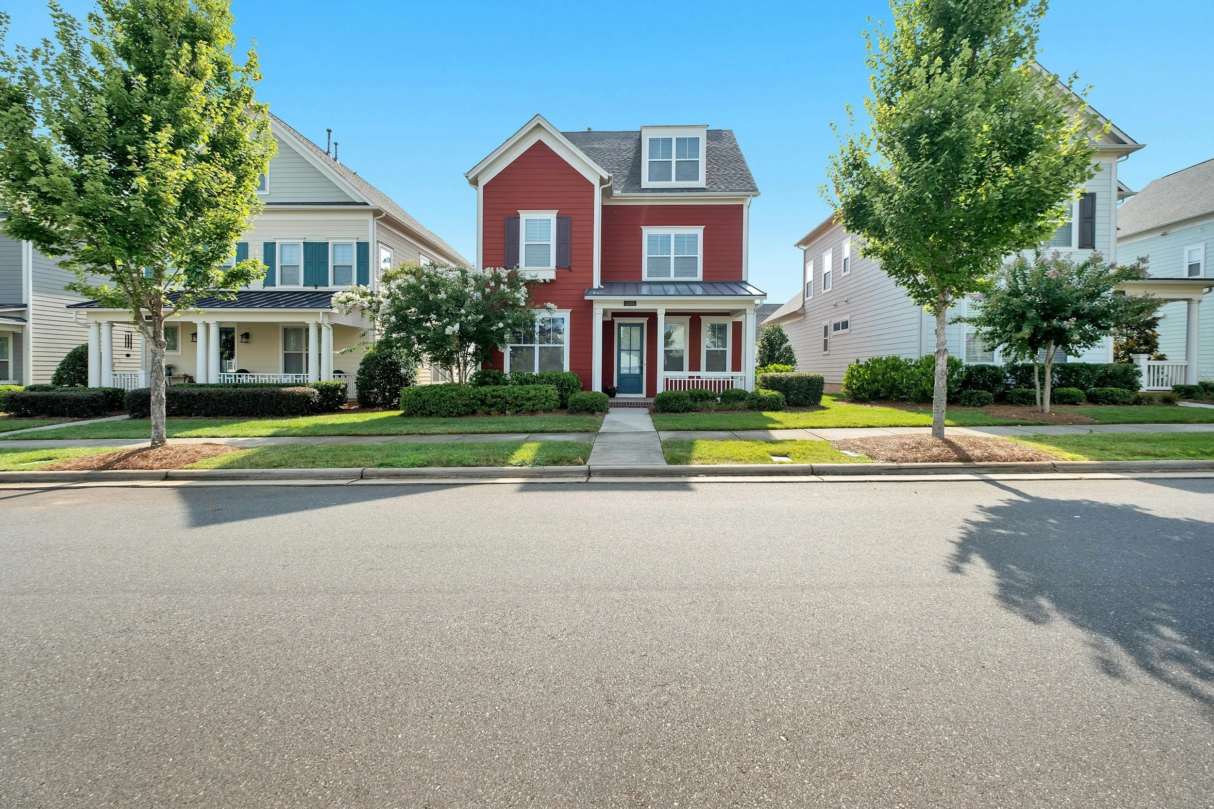Front view of a red three-story house with a porch, in a neighborhood with similar white houses, green trees, and a green lawn under a clear blue sky.