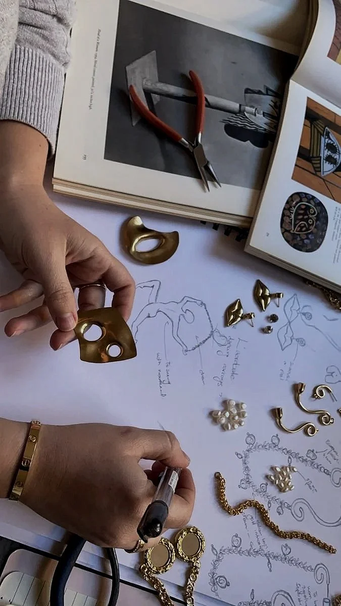 A workspace with jewelry designs, including gold earrings, a bracelet, pearl embellishments, and sketches. Tools like pliers and a pen are on the table, alongside open jewelry and art books.