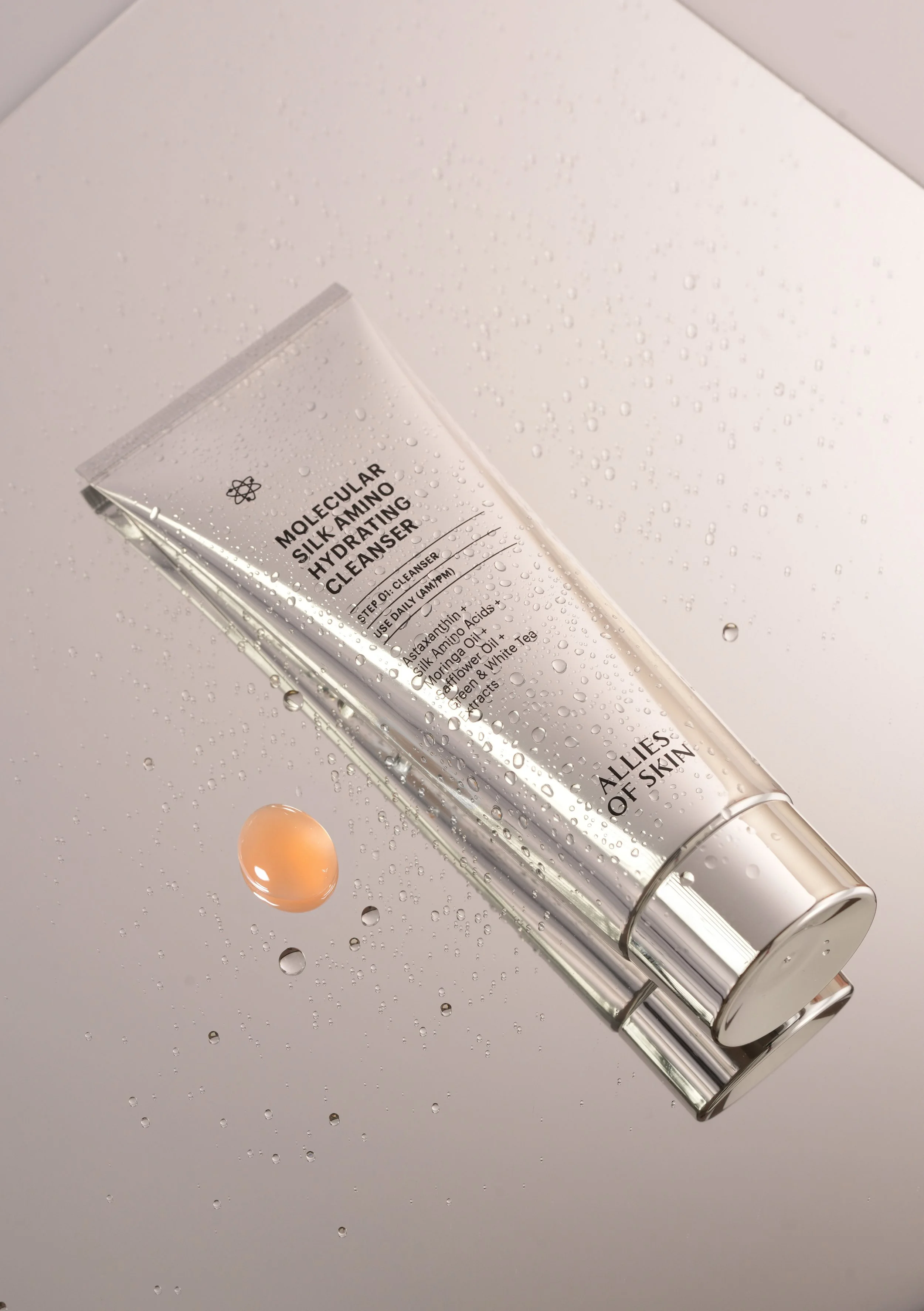 A silver tube of Molecular Silky Amo Cleaning product with water droplets on it, placed on a reflective surface with water droplets and a small drop of liquid in front.