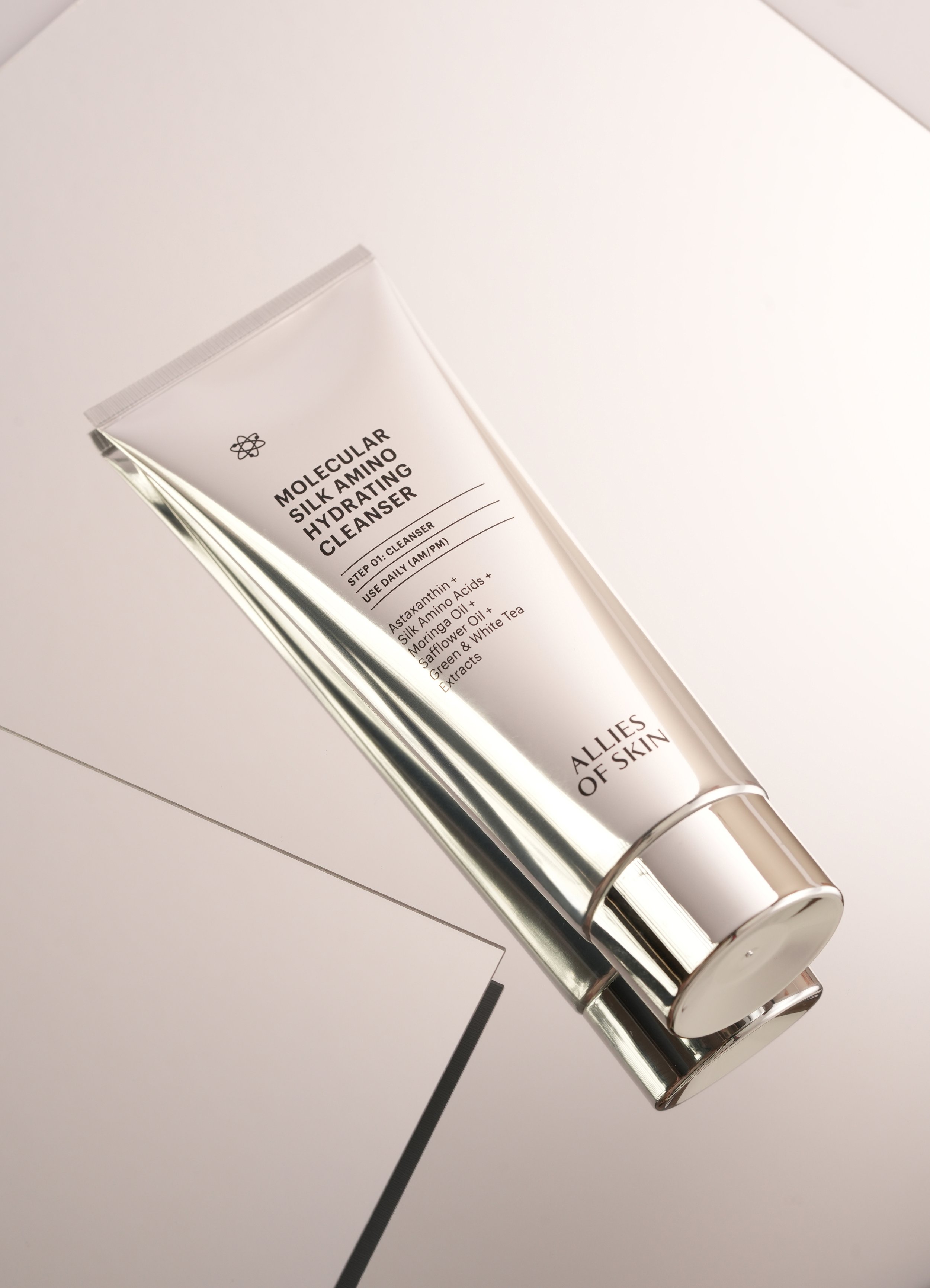 A silver tube of Molecular Silk Amino Hydrating Cleanser lying diagonally on a white surface with a reflective mirror beneath it.