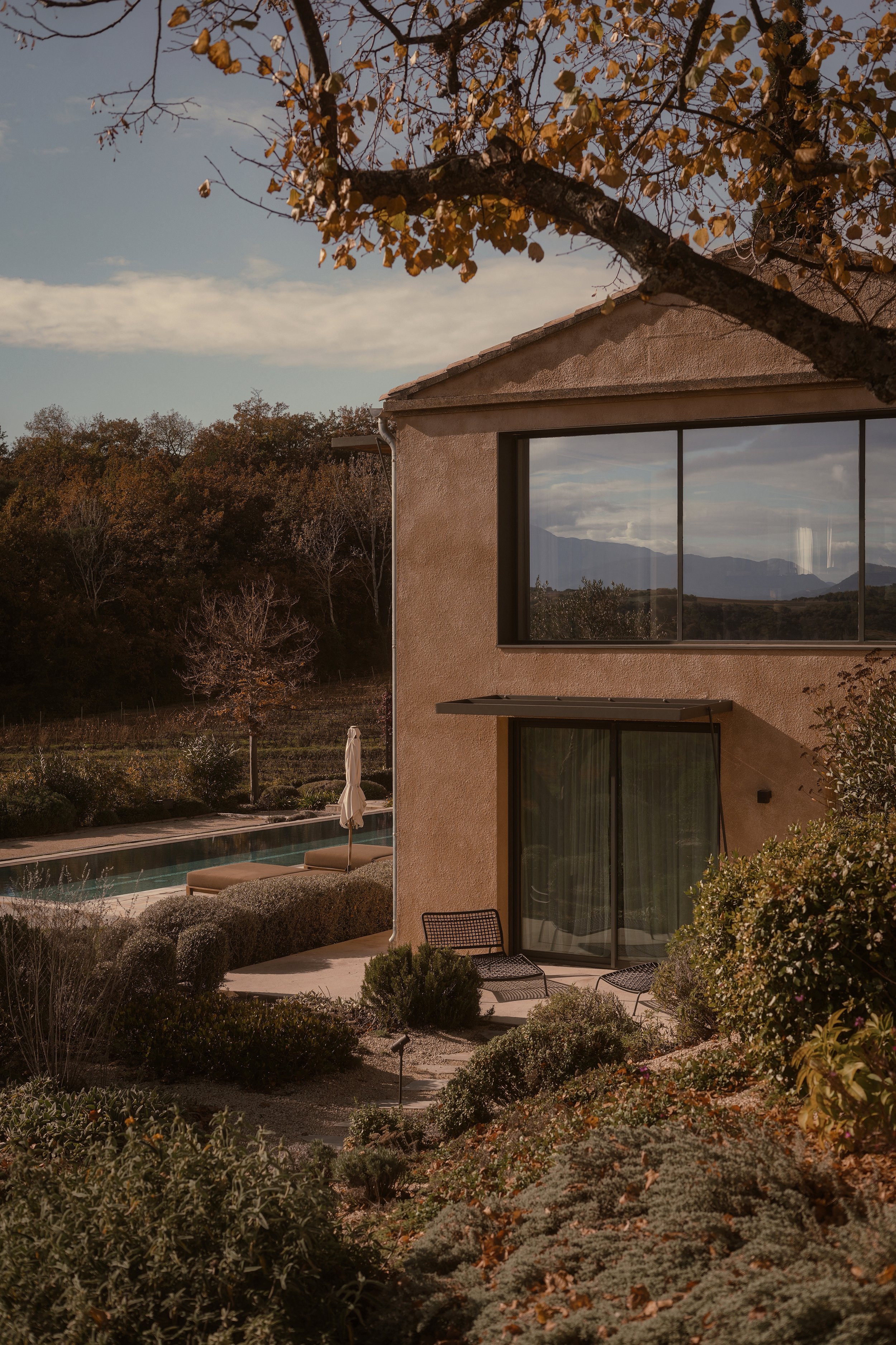 A modern two-story house with large windows, an outdoor patio with chairs and an umbrella, a swimming pool, and a landscaped garden in a rural area with mountains in the background, during autumn.
