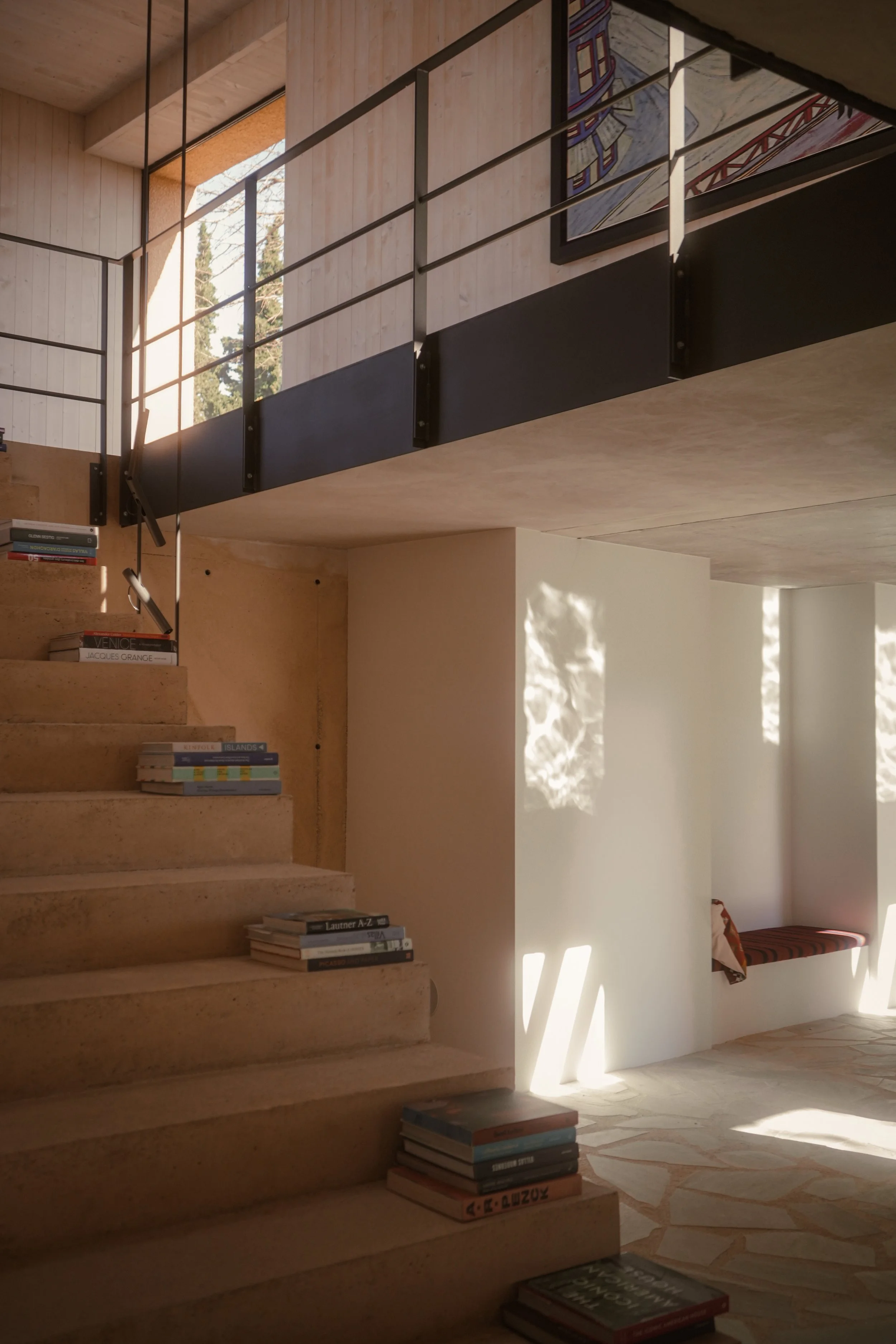 Interior view of a modern house with staircase, books on stairs and floor, sunlight streaming through windows, and a colorful painting on the wall.