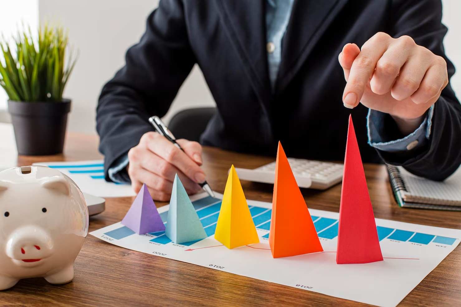Person in business attire pointing at color-coded 3D paper pyramids on a chart, with a piggy bank, calculator, and office supplies on the wooden desk.