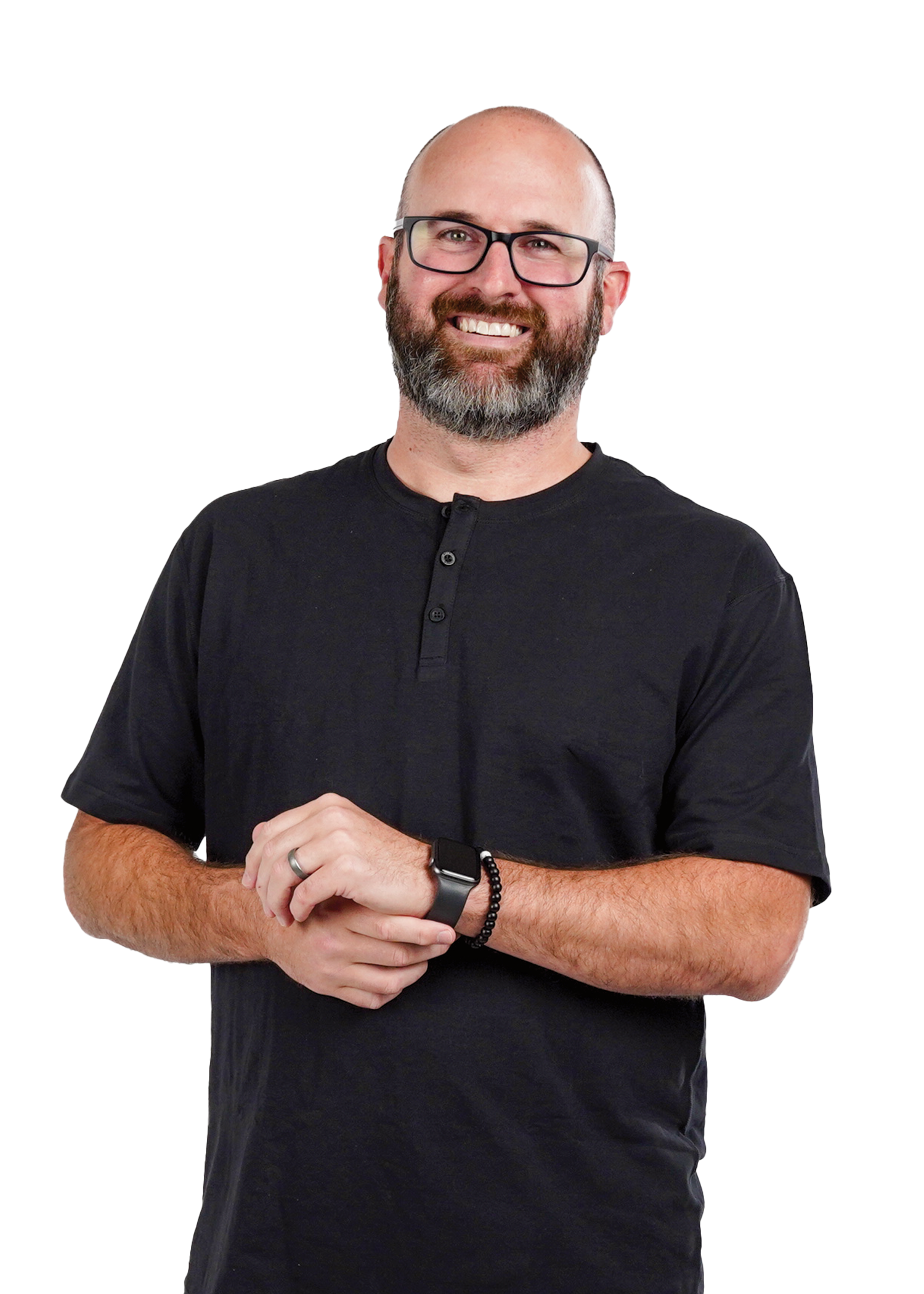 A smiling man with glasses, a beard, and a bald head, wearing a black short-sleeve shirt, a watch, and a beaded bracelet, standing with hands clasped.