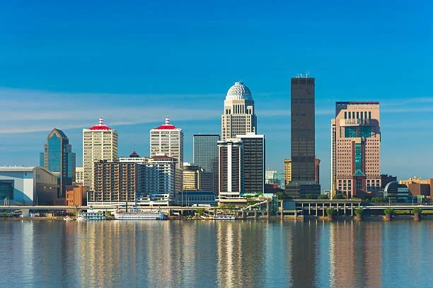 View of a city skyline with tall buildings reflected in a river under a clear blue sky.